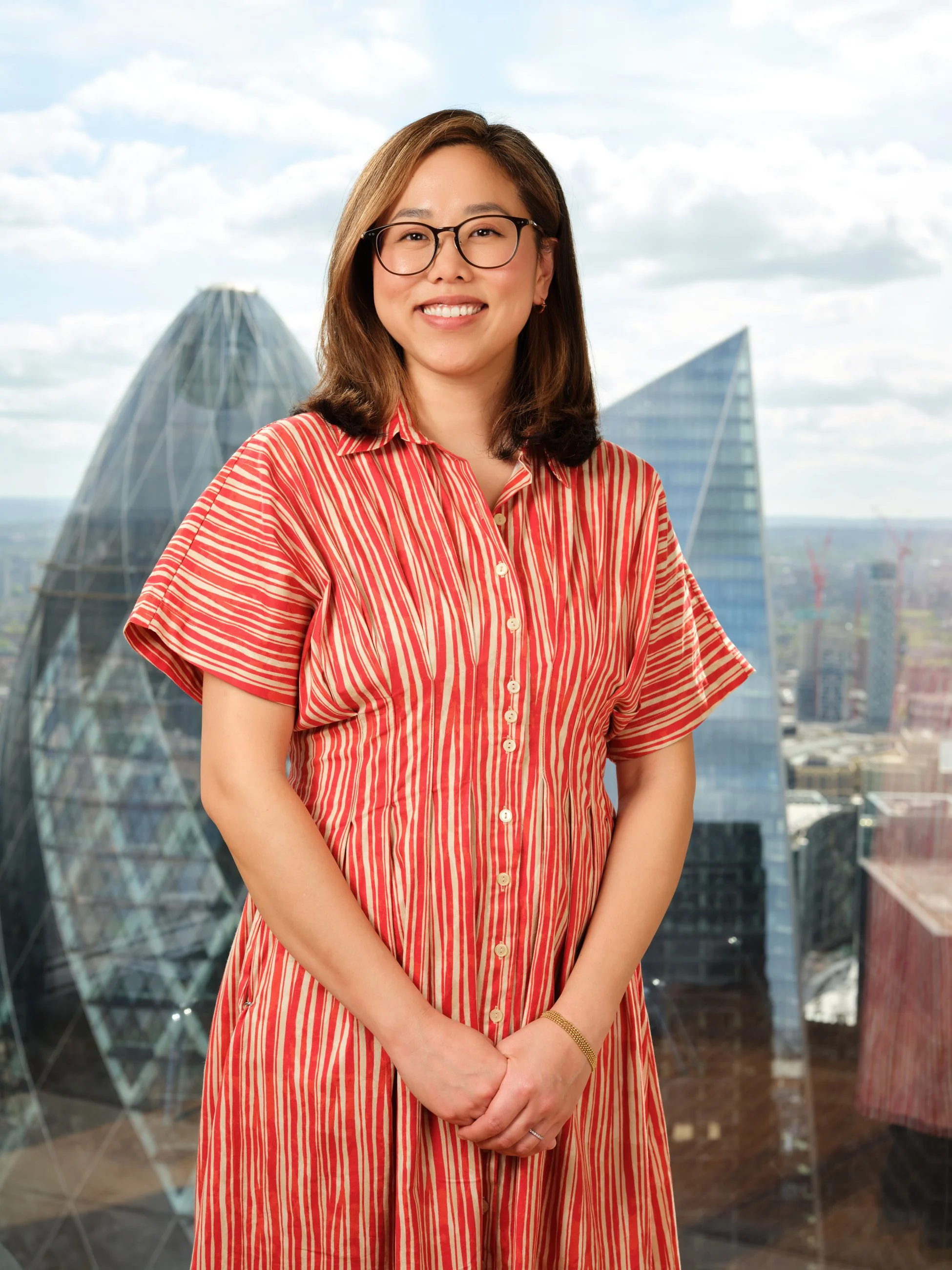 A woman with glasses and shoulder-length hair standing in front of a cityscape with modern glass buildings, wearing a red and beige striped dress, smiling at the camera.
