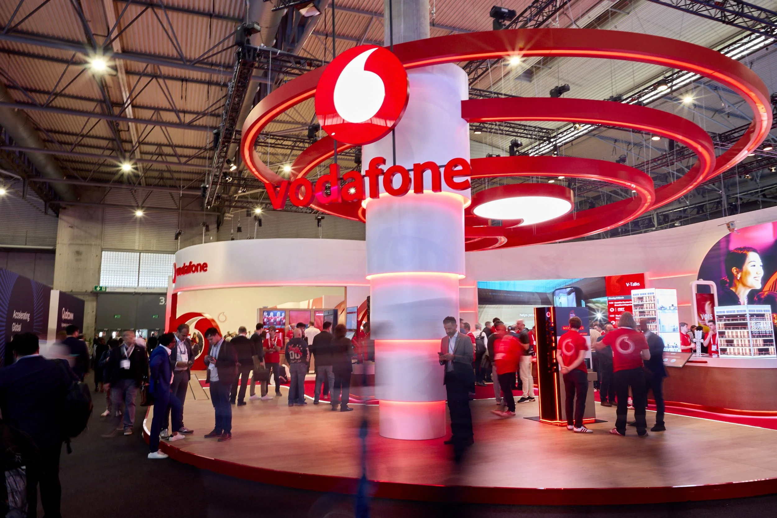 Vodafone booth at a technology exhibition, featuring a large red circular logo and numerous visitors interacting with displays.