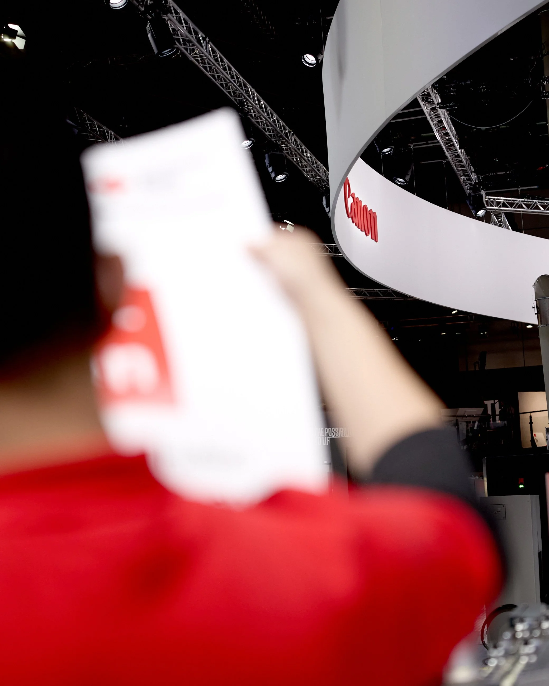 Blurred person holding a white object in front of a large Canon sign at an indoor convention or event