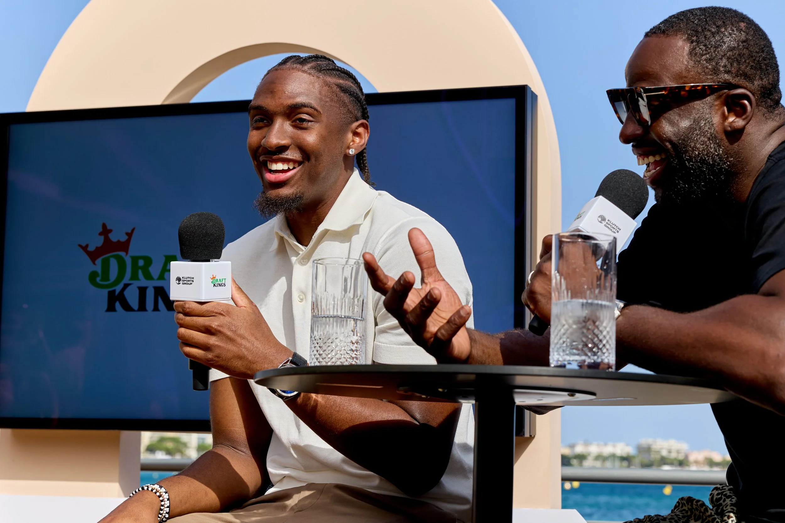 Two men sitting at a table with microphones and glasses of water, speaking during a televised event with a large screen behind them that says Draft Kings.