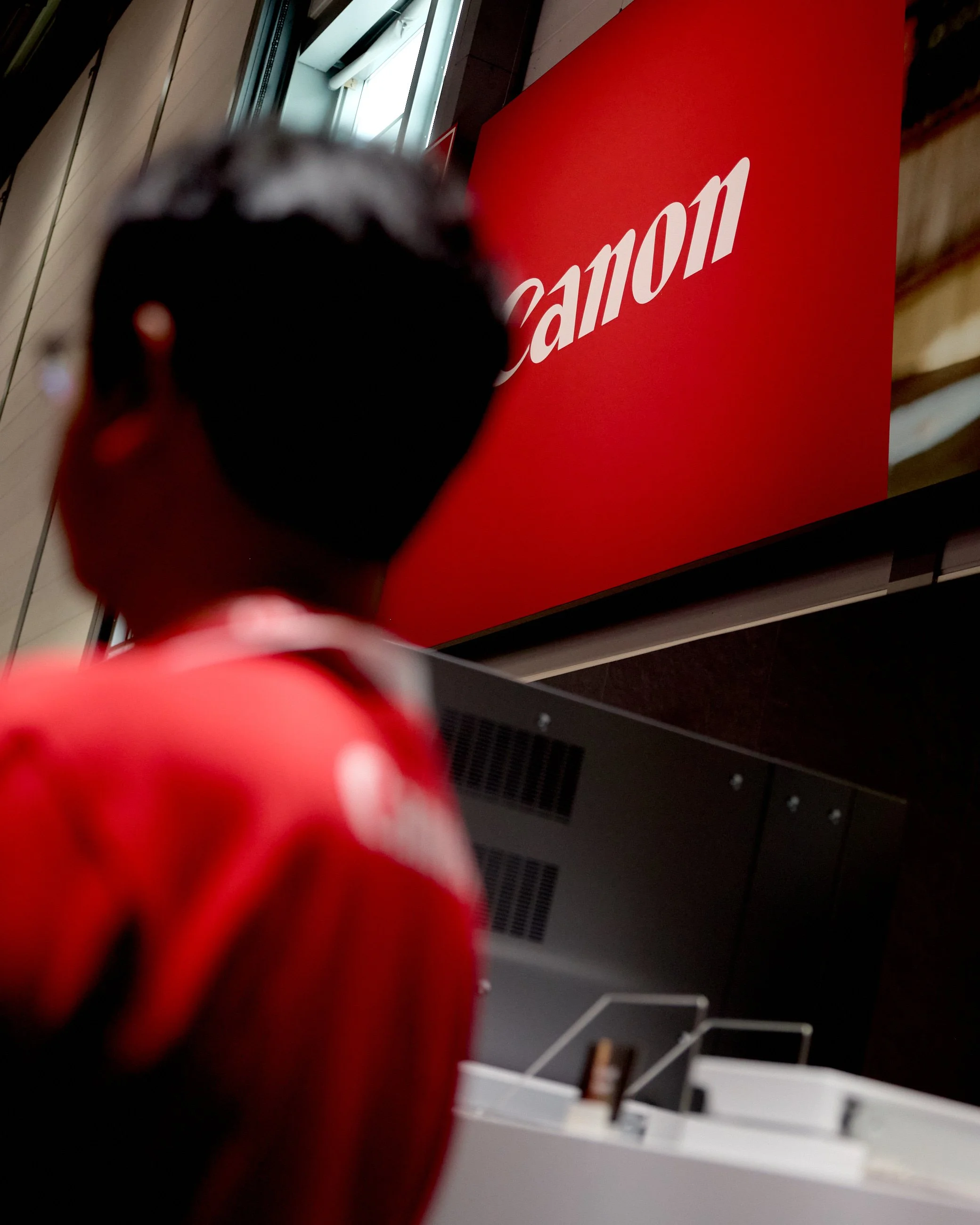 Blurred close-up of a person standing in front of a large Canon sign at an indoor location.