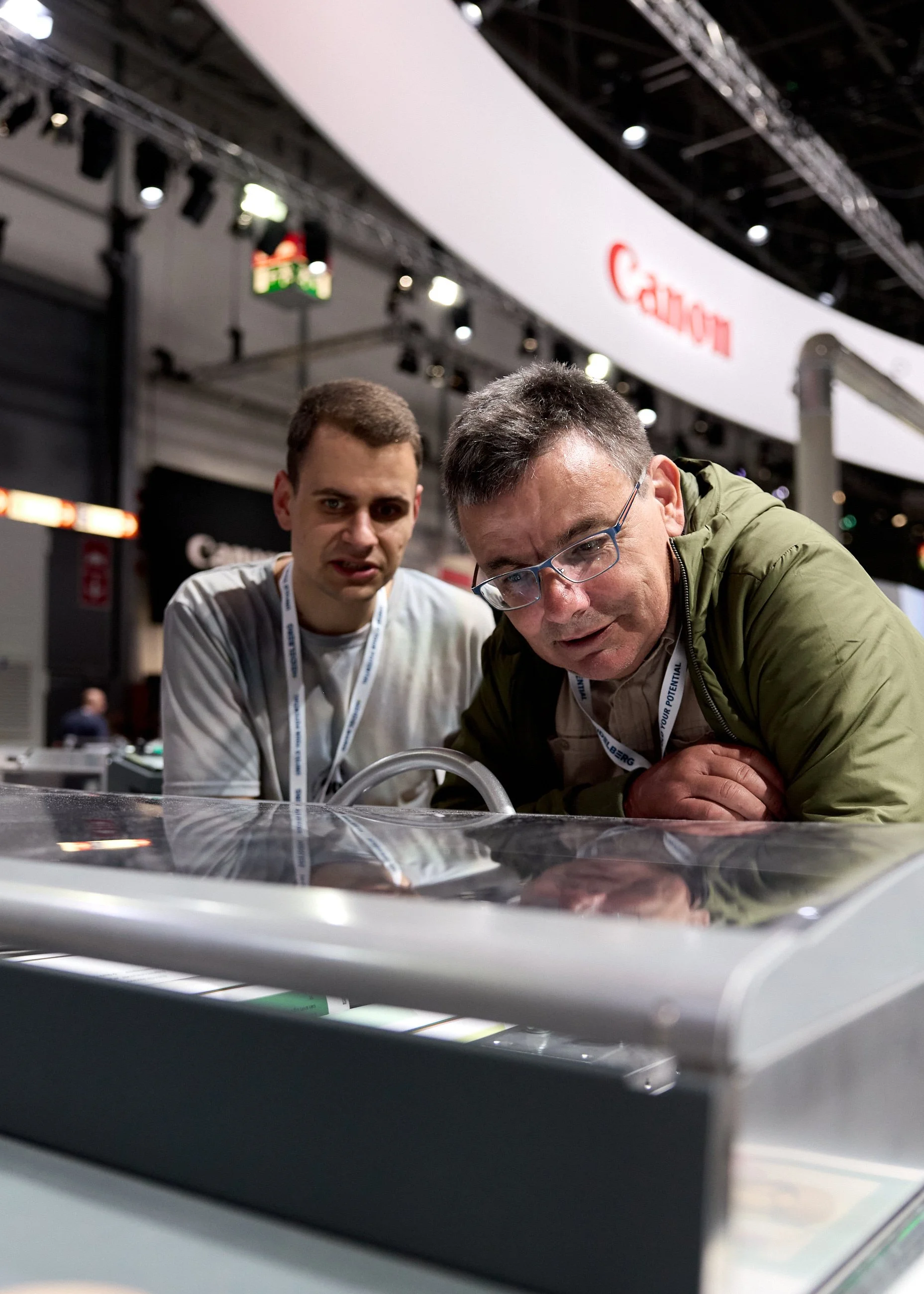 Two men examining a display or machine at a technology exhibition, with Canon branding in the background.