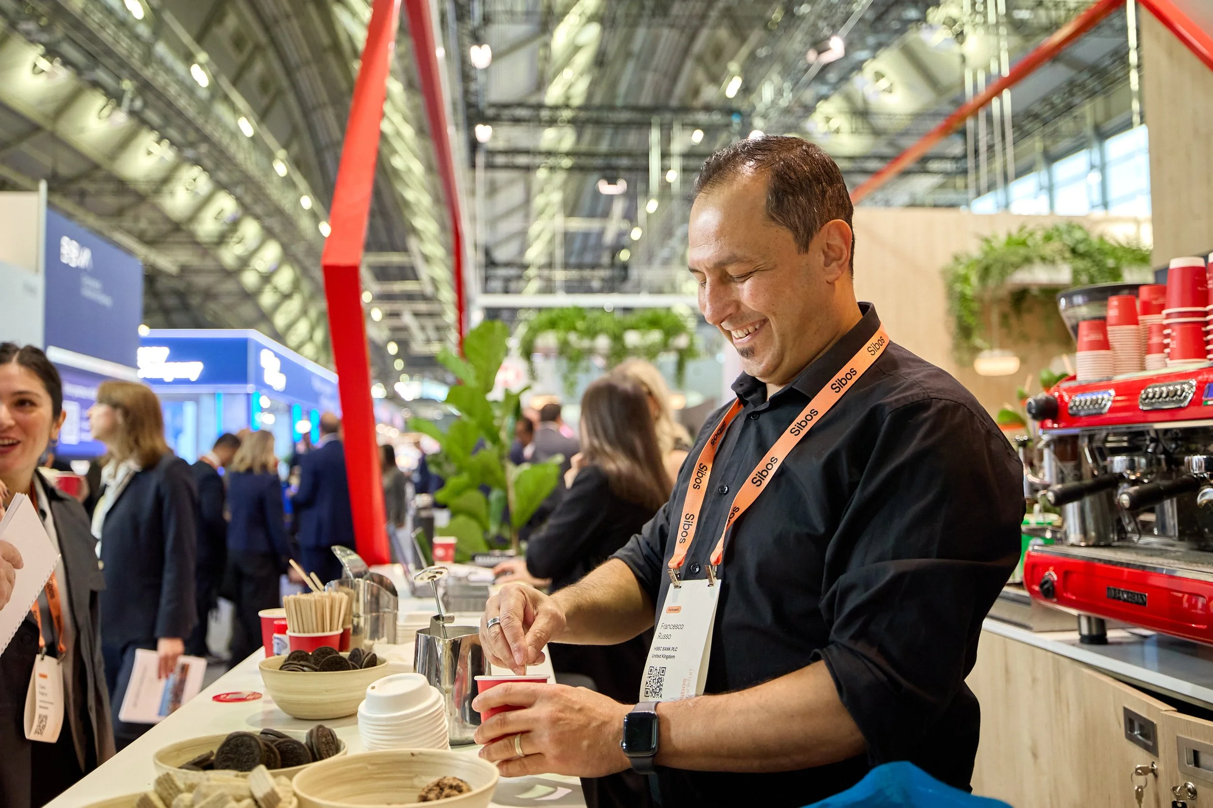 A man with a black shirt and orange lanyard, smiling while preparing a drink at a coffee booth during a conference or trade show.
