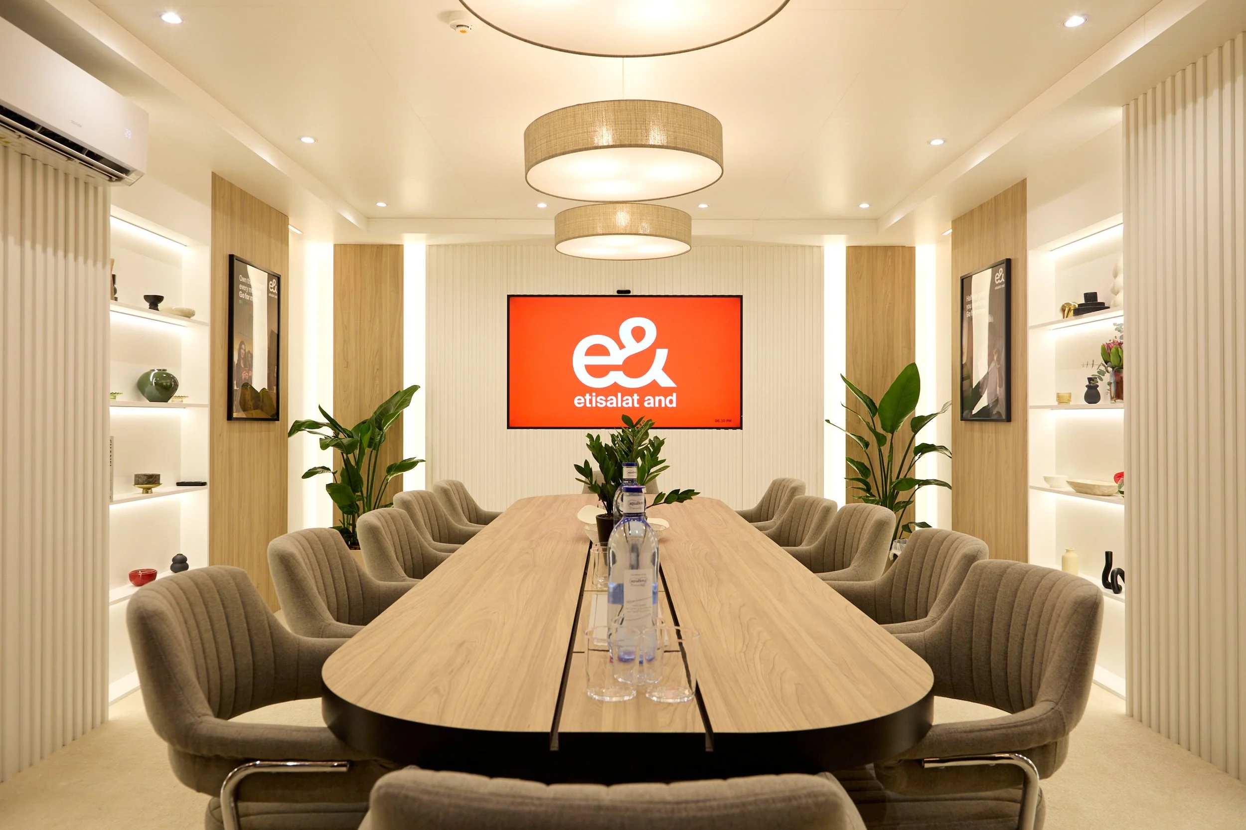 Modern conference room with a long wooden table surrounded by beige upholstered chairs, indoor plants, and decorative shelves with vases and art pieces. A large red screen displays the 'etisalat and' logo.