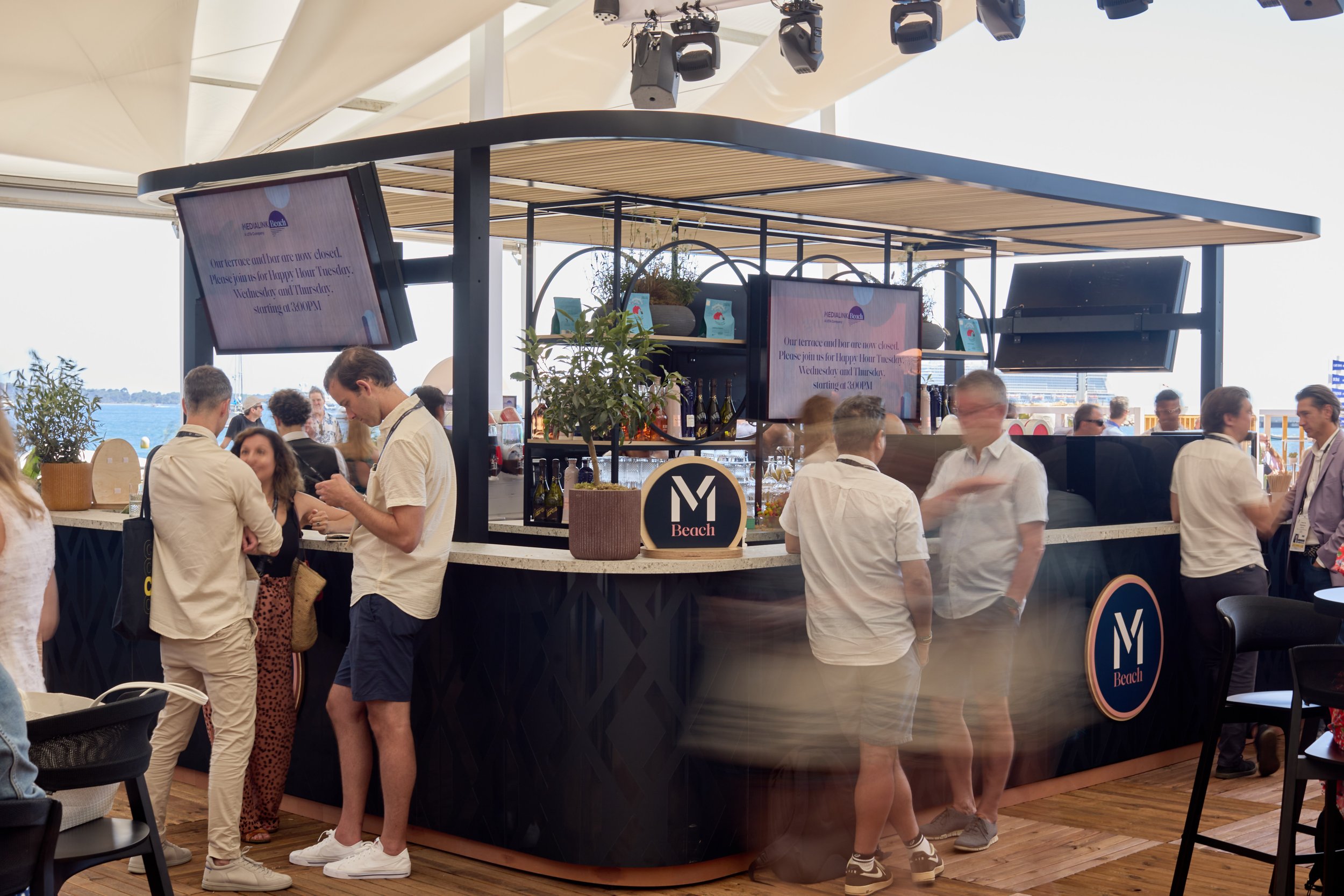 A bar area on a boat or yacht with people socializing and drinking, under a canopy with two TV screens displaying information about the hour and channel, and ocean visible in the background.