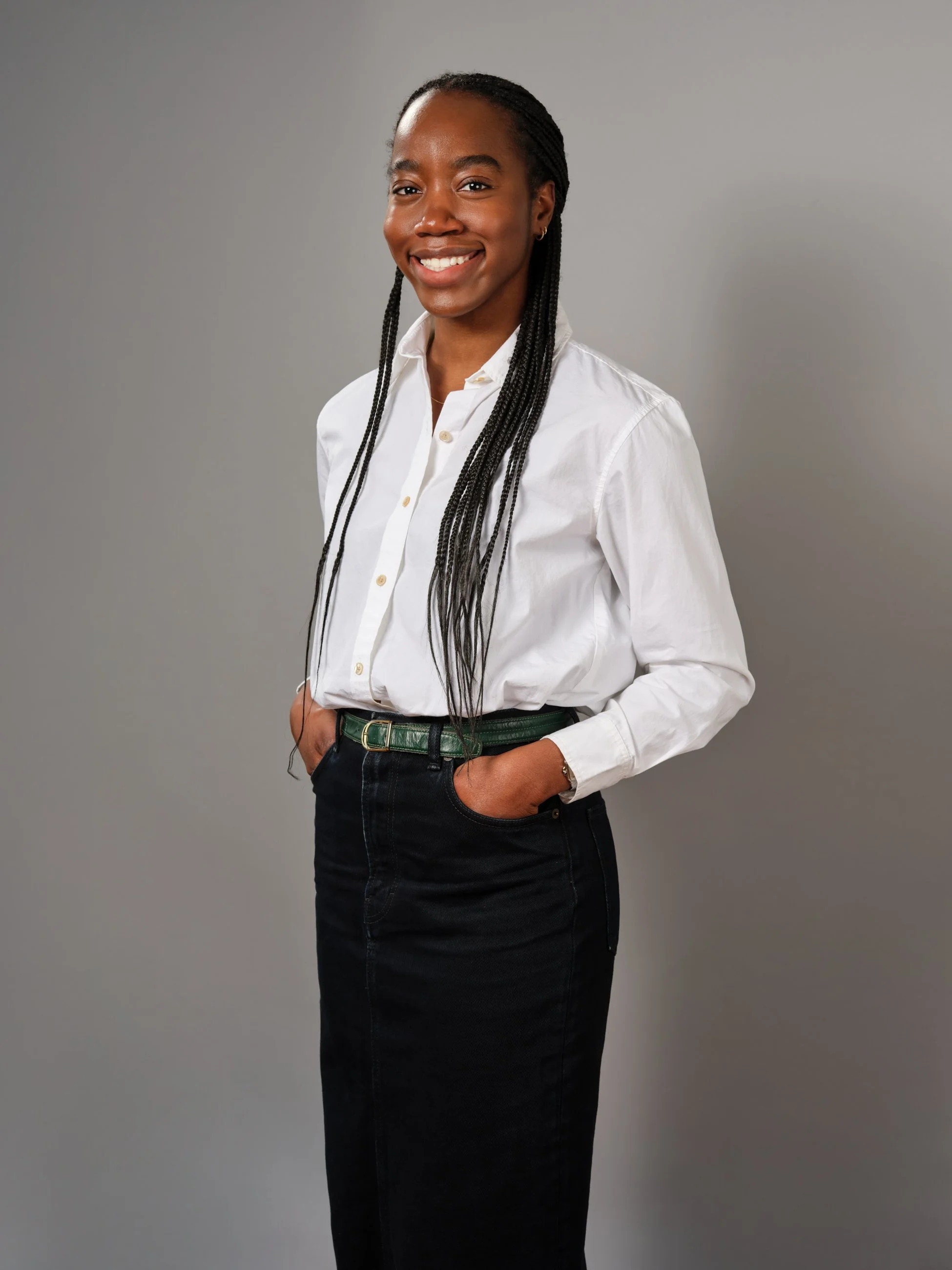 A smiling woman with long braided hair wearing a white button-up shirt and a dark skirt, standing with hands in pockets on a plain gray background.