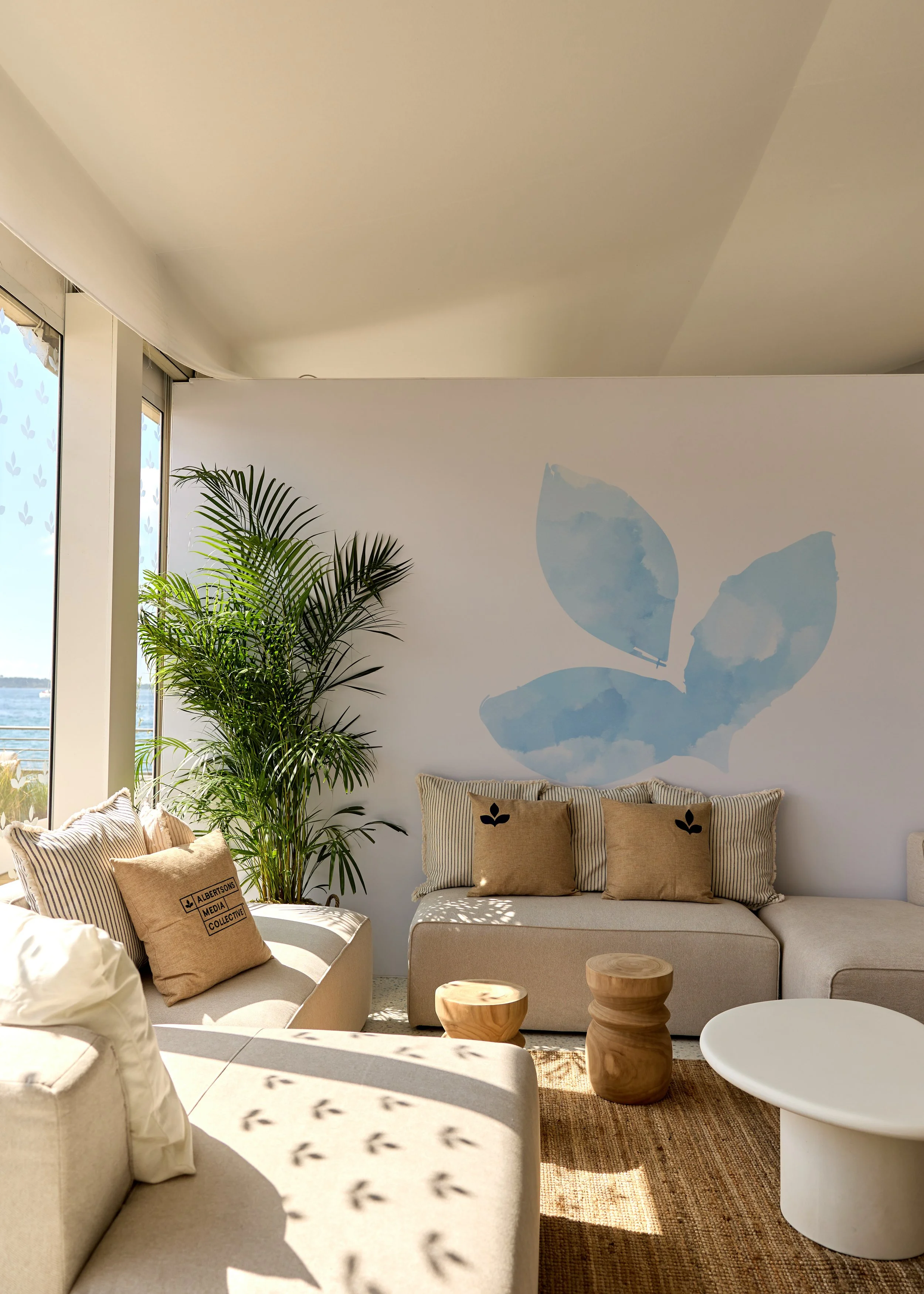Modern beachside living room with beige sofas, striped throw pillows, a large potted plant, and a watercolor butterfly mural on the white wall, sunlight streaming through large windows.