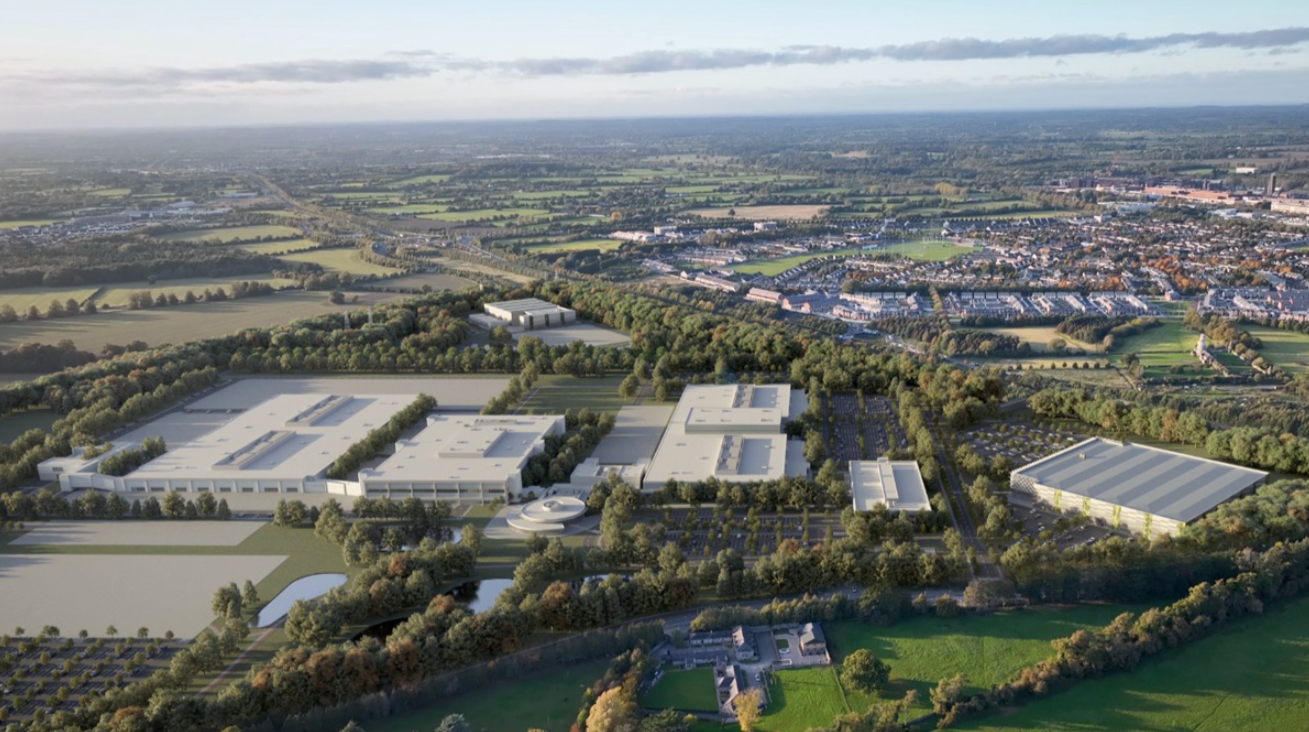 Aerial view of Kildare Innovation Campus, multiple buildings surrounded by green spaces, trees, and parking lots, extending into a suburban area with neighbourhoods and open fields in the distance.