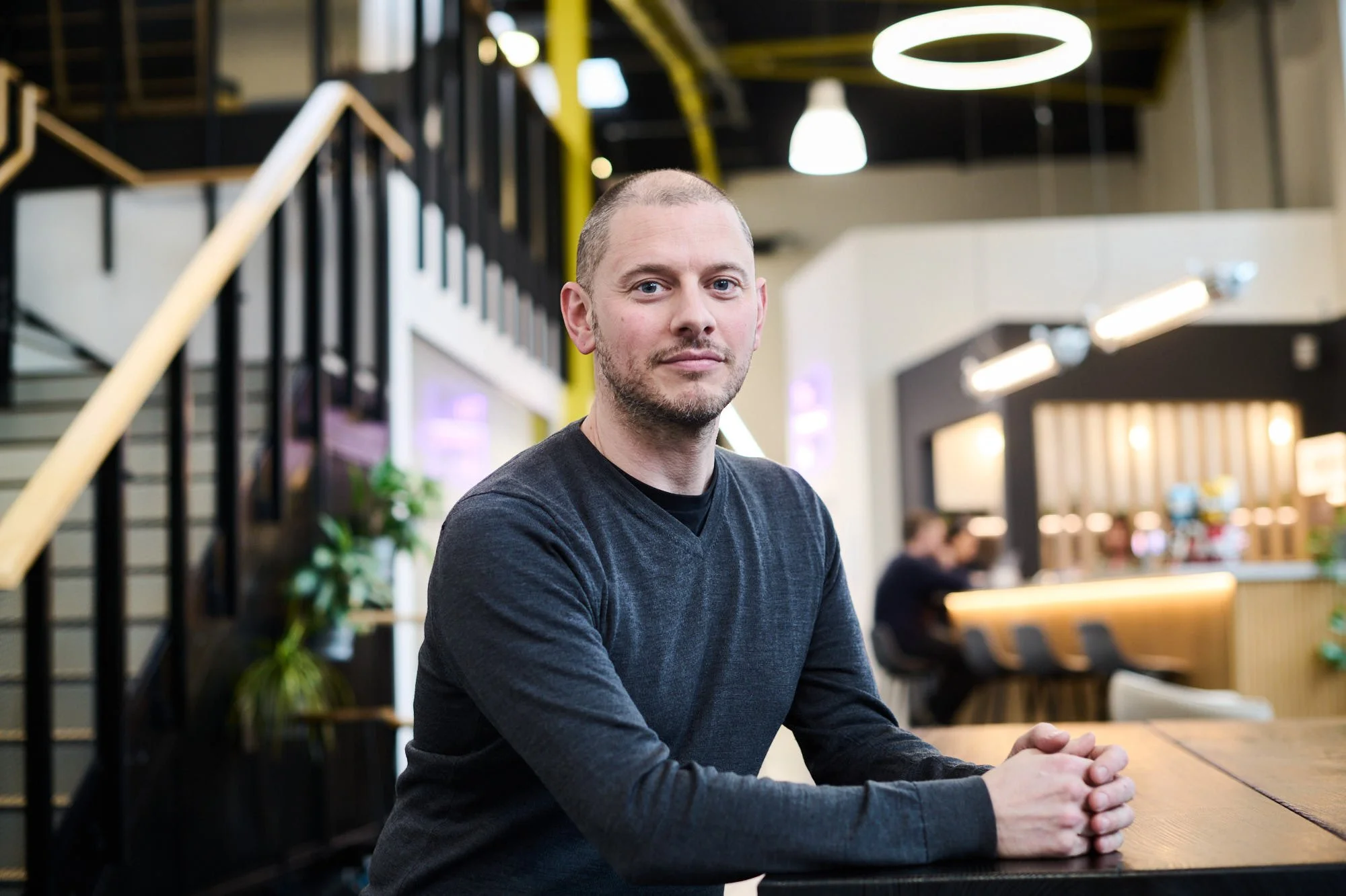 Dr Ashley Brewer, Head of Incubators at Science Creates. A man with short hair and a beard sitting at a table in a modern, brightly lit Deep Tech incubator atrium space and bar, Science Creates Old Market.