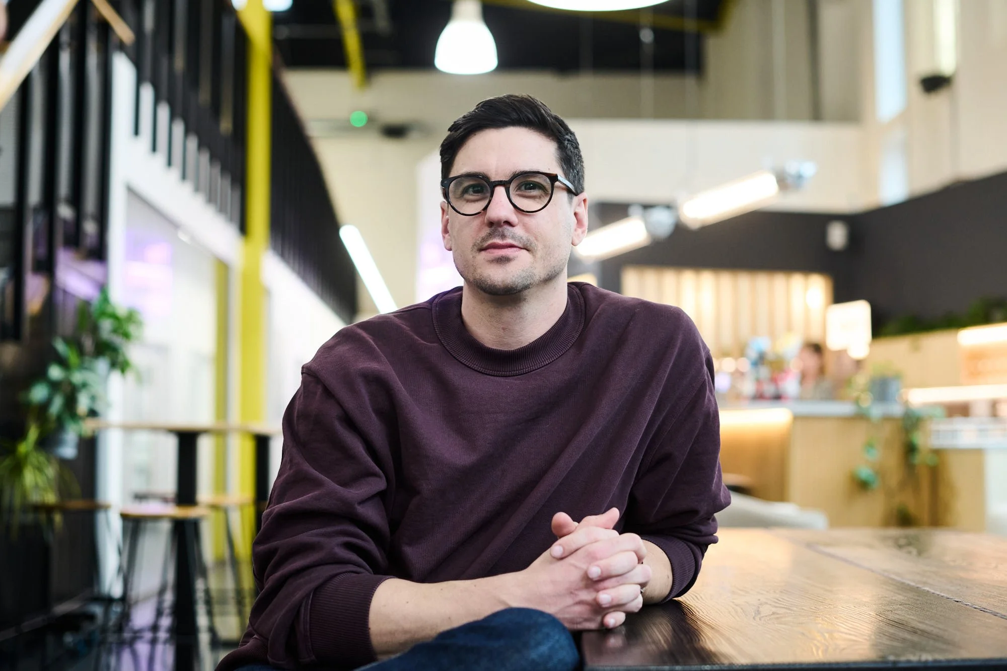 Dr Harry Destecroix, Executive Chair of Science Creates. A man with dark hair and glasses sitting at a wooden table in a modern, brightly lit Deep Tech incubator atrium space and bar, Science Creates Old Market.