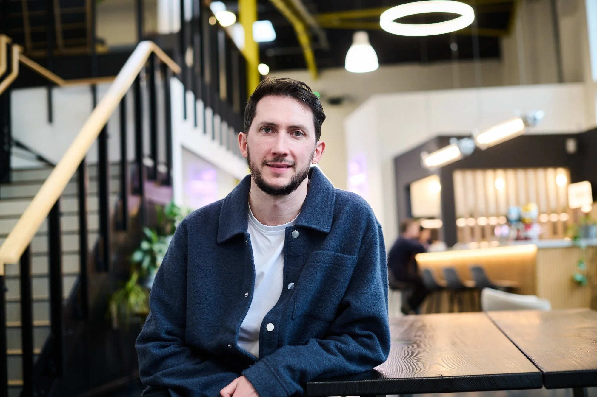 Patrick Fallon MArch ARB, Head of Development at Science Creates. A man sitting at a wooden table in a modern, well-lit Deep Tech incubator atrium space and bar, Science Creates Old Market.