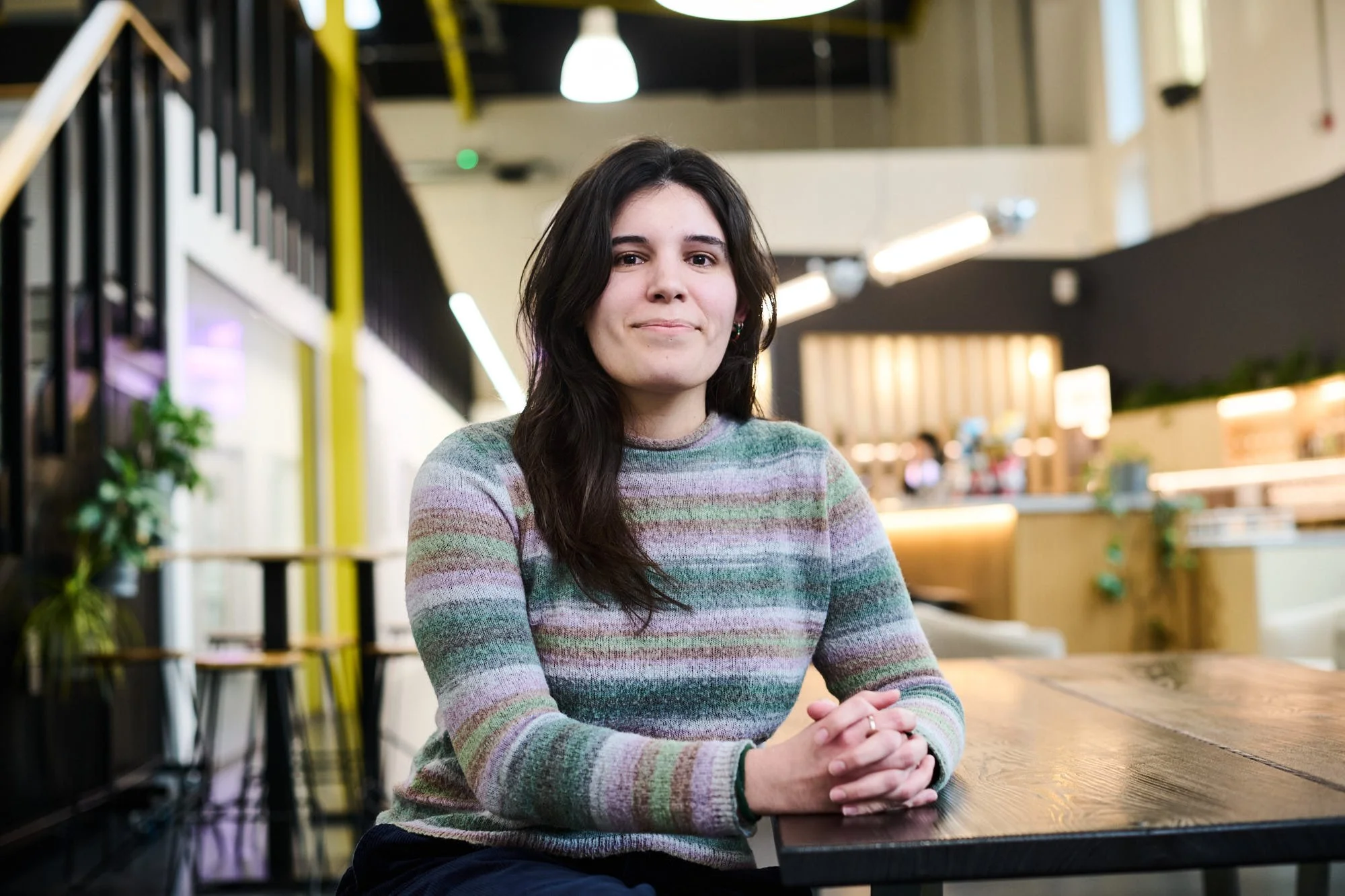 Irene Pastor MArch ARB, Architect at Science Creates. A woman with long dark hair sitting at a wooden table in a modern, well-lit Deep Tech incubator atrium space and bar, Science Creates Old Market.