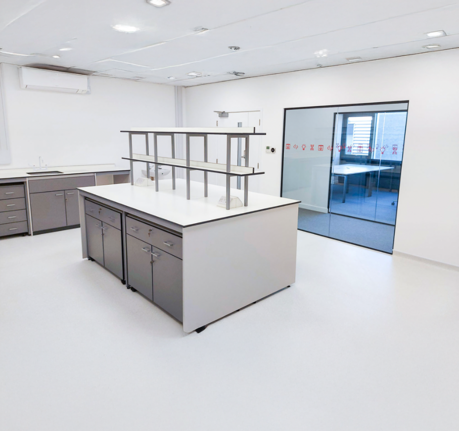 Singer Instruments R&D Lab with white walls and flooring, equipped with a central worktable that has grey cabinets and a metal railing on top. There is a room with glass doors and red safety symbols on the glass to the right.