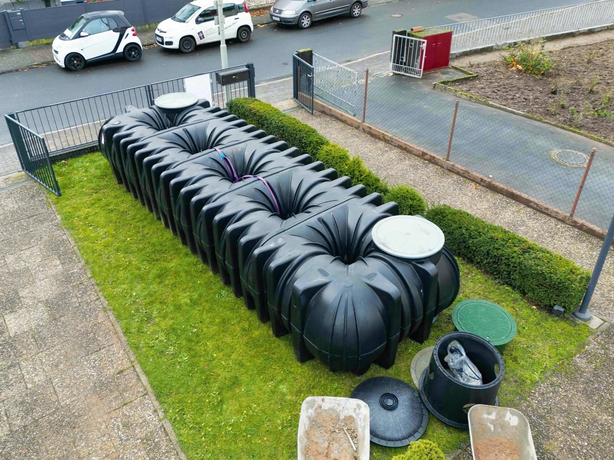 Mehrere schwarze Generatortanks und das Zubehör sind auf einem kleinen Rasenstück im Freien. Es gibt auch eine grüne und eine schwarze Abdeckung, sowie einige Mülltonnen und Behälter. Im Hintergrund ist eine Straße mit parkenden Autos.