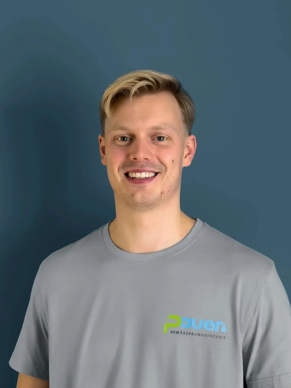 Ein junger Mann trägt ein graues T-Shirt mit einem Logo der Firma 2Power Wassertechnik und lächelt vor einem blauen Hintergrund.