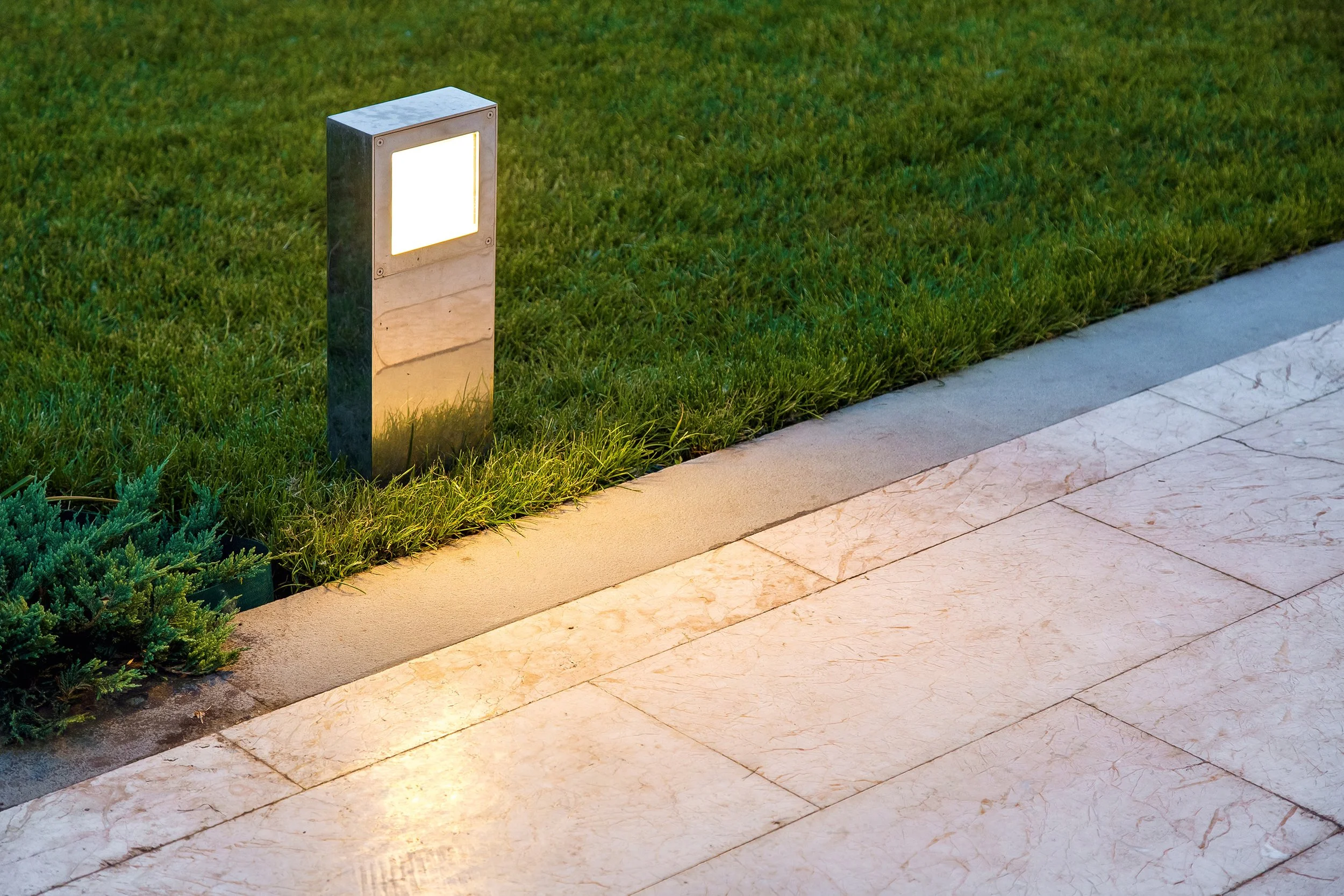 Ein beleuchteter Wegleuchte an einem Rasenstreifen neben einem Gehweg aus Marmorplatten, bei Dämmerung oder Nacht.