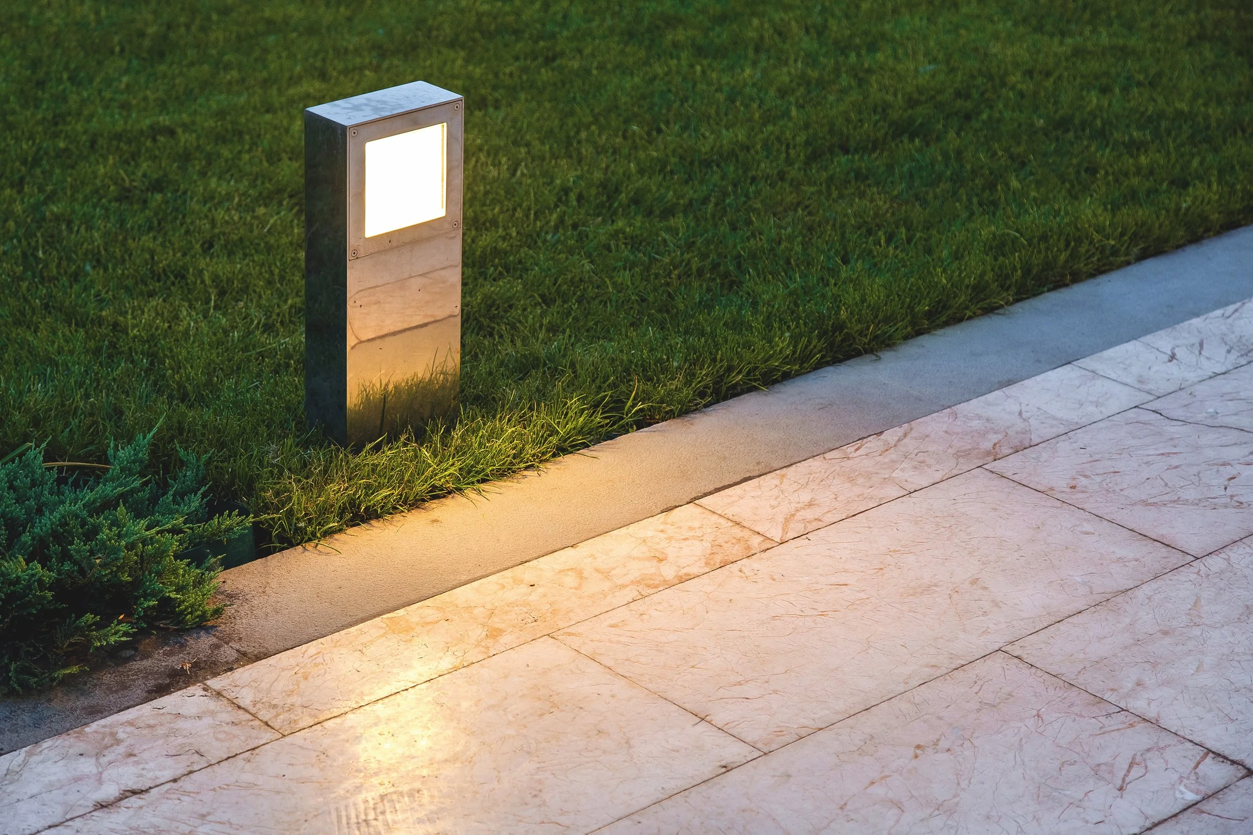 Ein beleuchteter Wegleuchten am Rande eines Gehwegs neben grünem Rasen und kleinen Pflanzen, nachts.