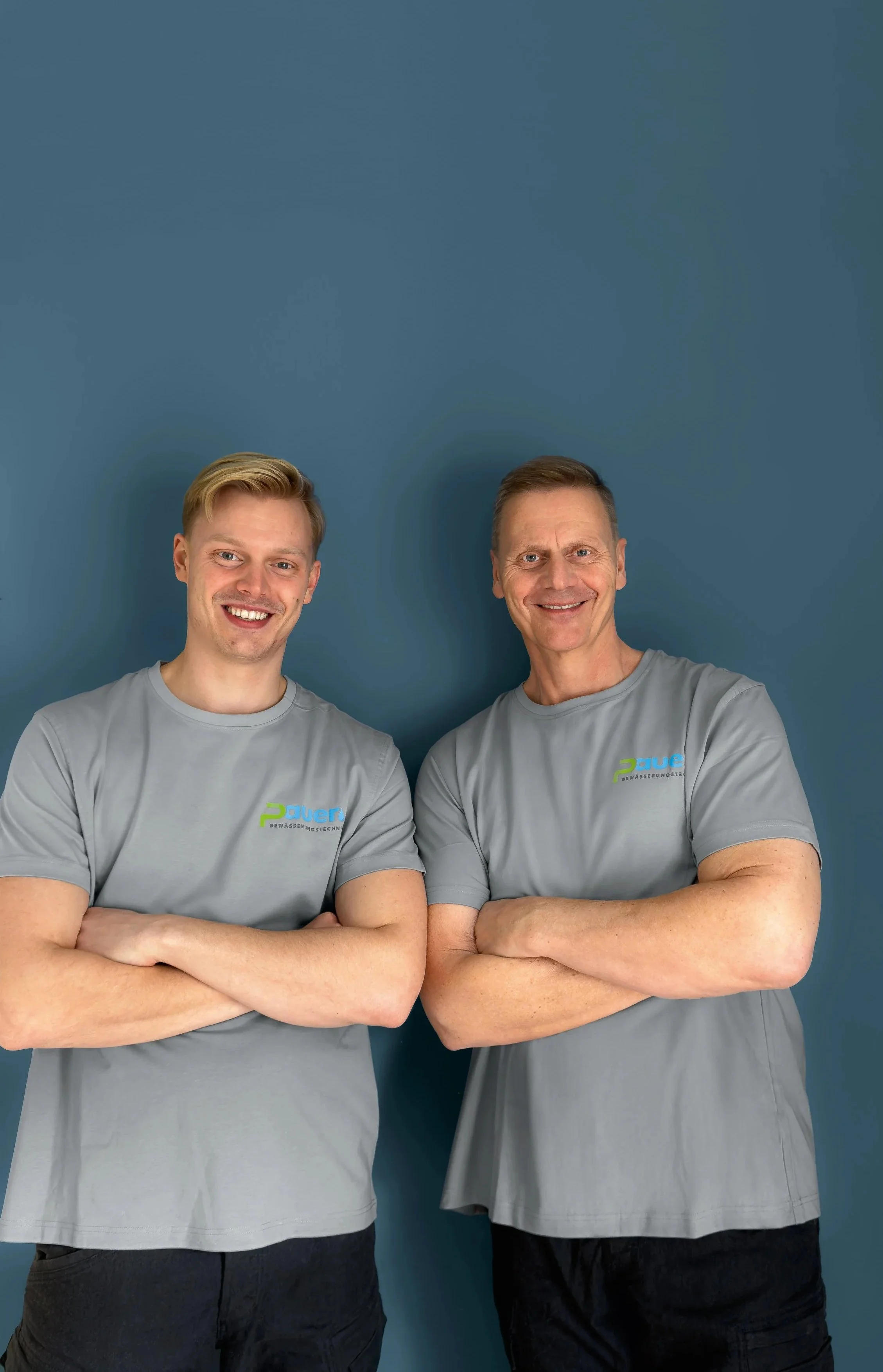 Zwei Männer in grauen T-Shirts mit geprägtem Logo, die vor einer blauen Wand stehen und die Arme verschränkt haben.