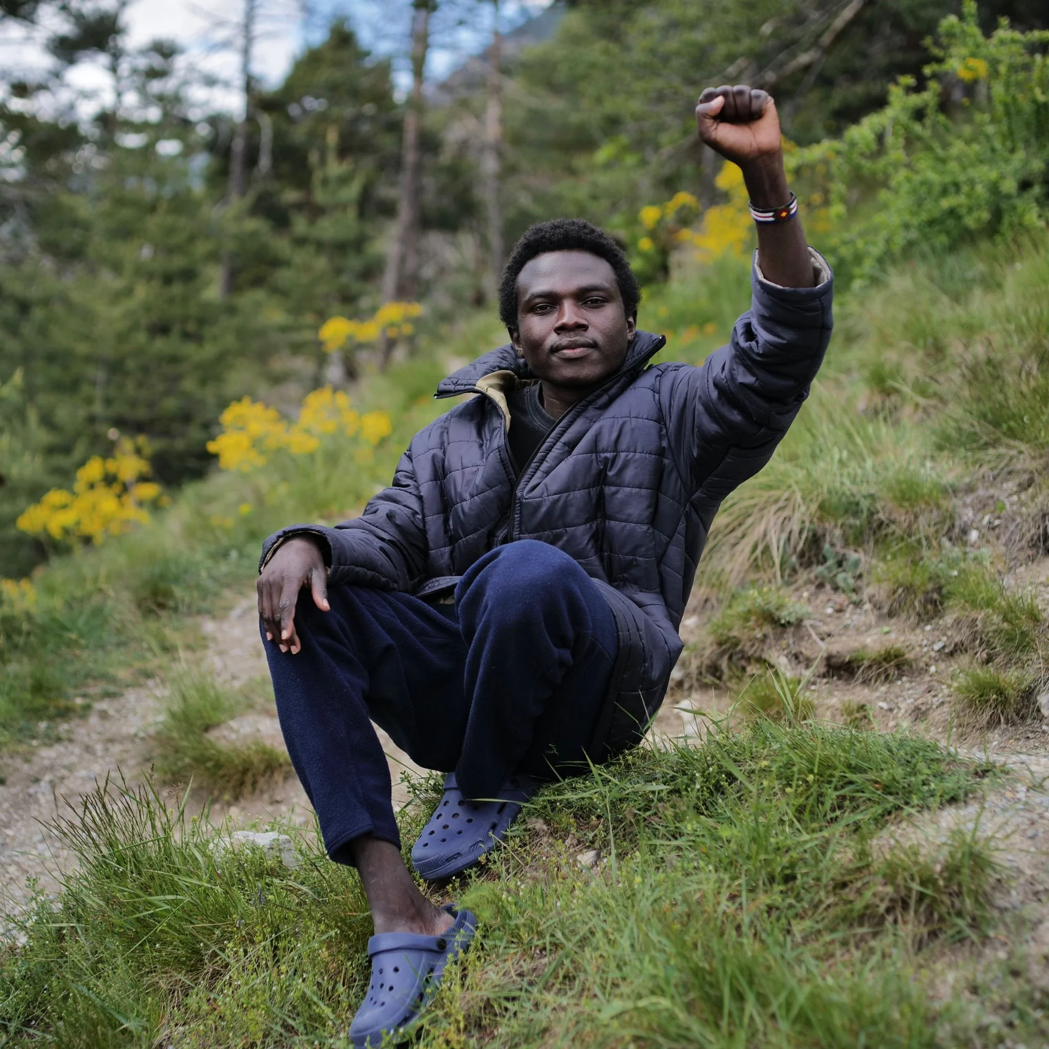 A young man with dark skin and curly hair crouches on a grassy hillside with yellow flowers, wearing a dark puffer jacket, dark pants, and blue Crocs, raising his right fist in a gesture of solidarity or strength amidst a green outdoor setting.