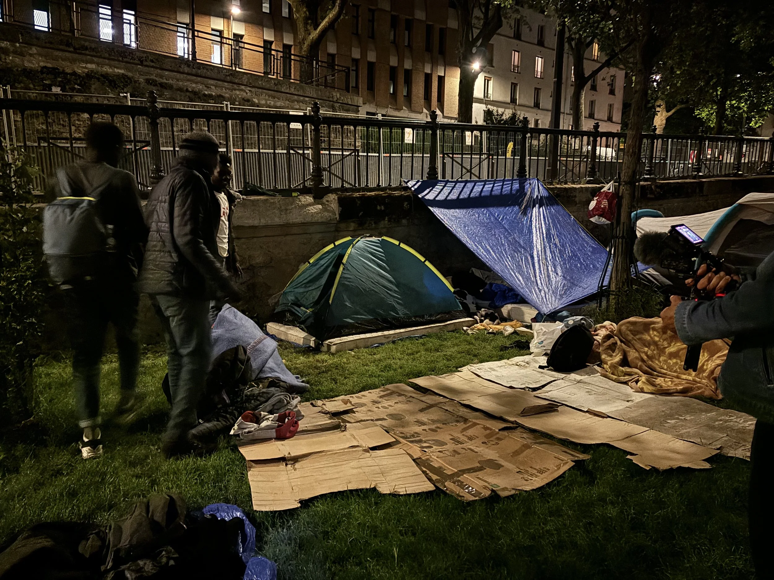 Nighttime scene of a small homeless encampment with tents, cardboard bedding, and people standing nearby.