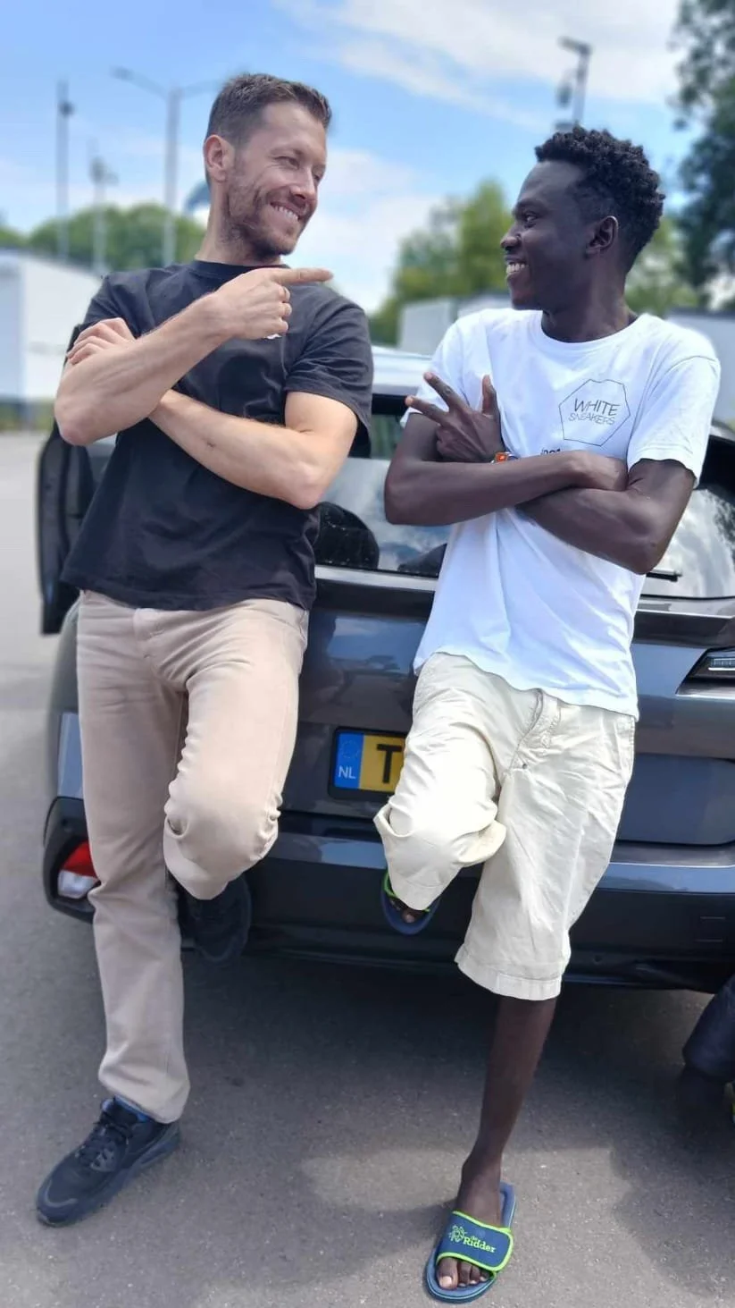 Two men are leaning against a car, smiling and engaging in conversation. One man is wearing a black T-shirt and beige pants, the other is wearing a white T-shirt and white shorts.