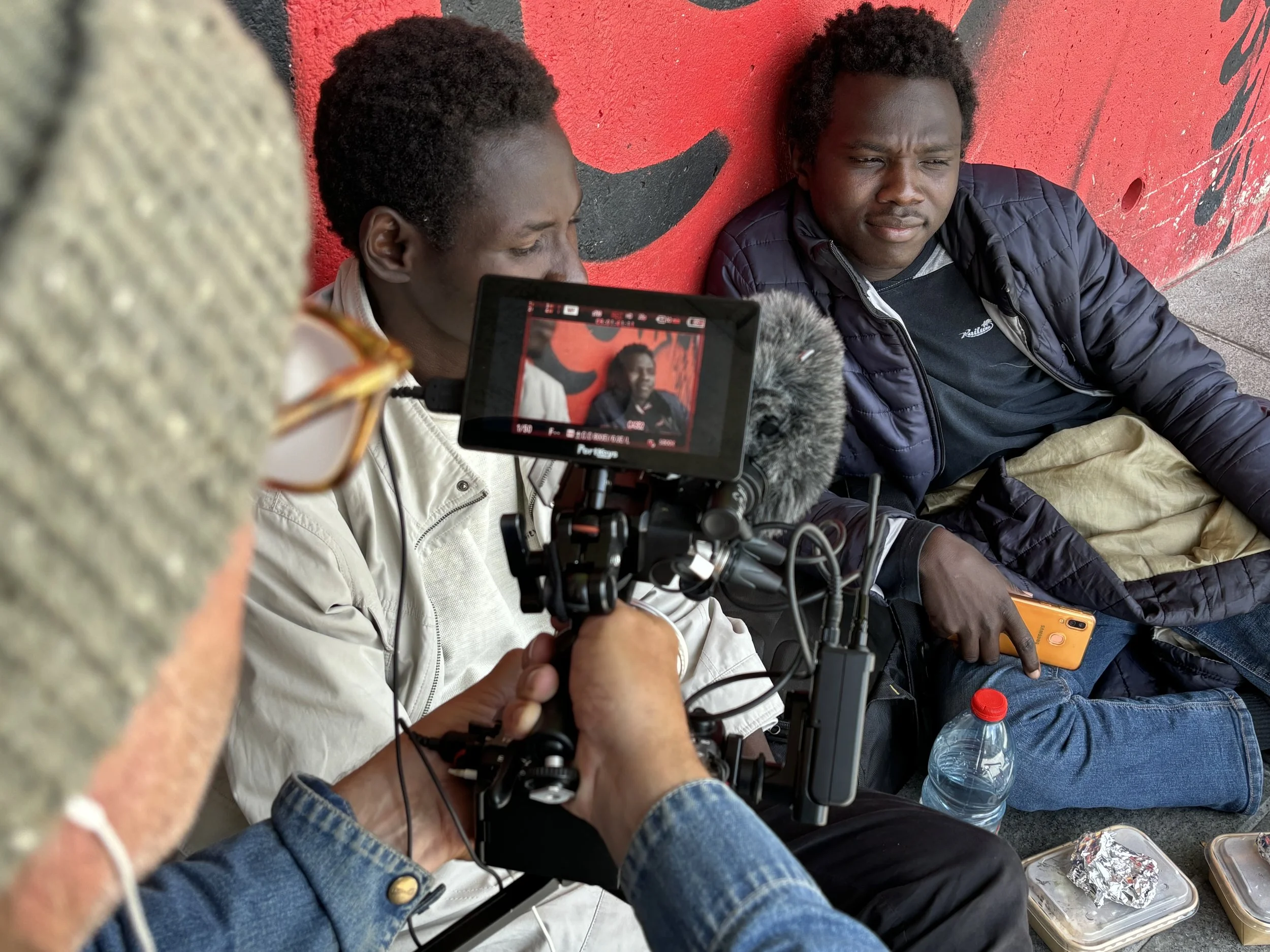 Two men sitting on the sidewalk against a red wall; one is being filmed with a camera while the other is holding a phone and looking serious.