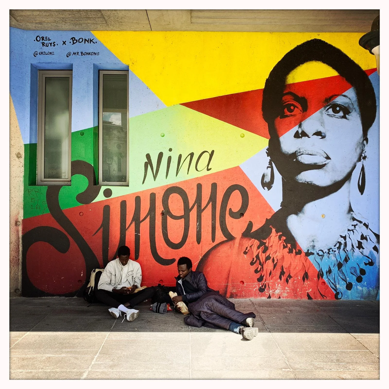 Two men sitting on the sidewalk with their heads down in front of a large, colorful mural of Nina Simone's face and the words 'Nina Simone' written on the wall.
