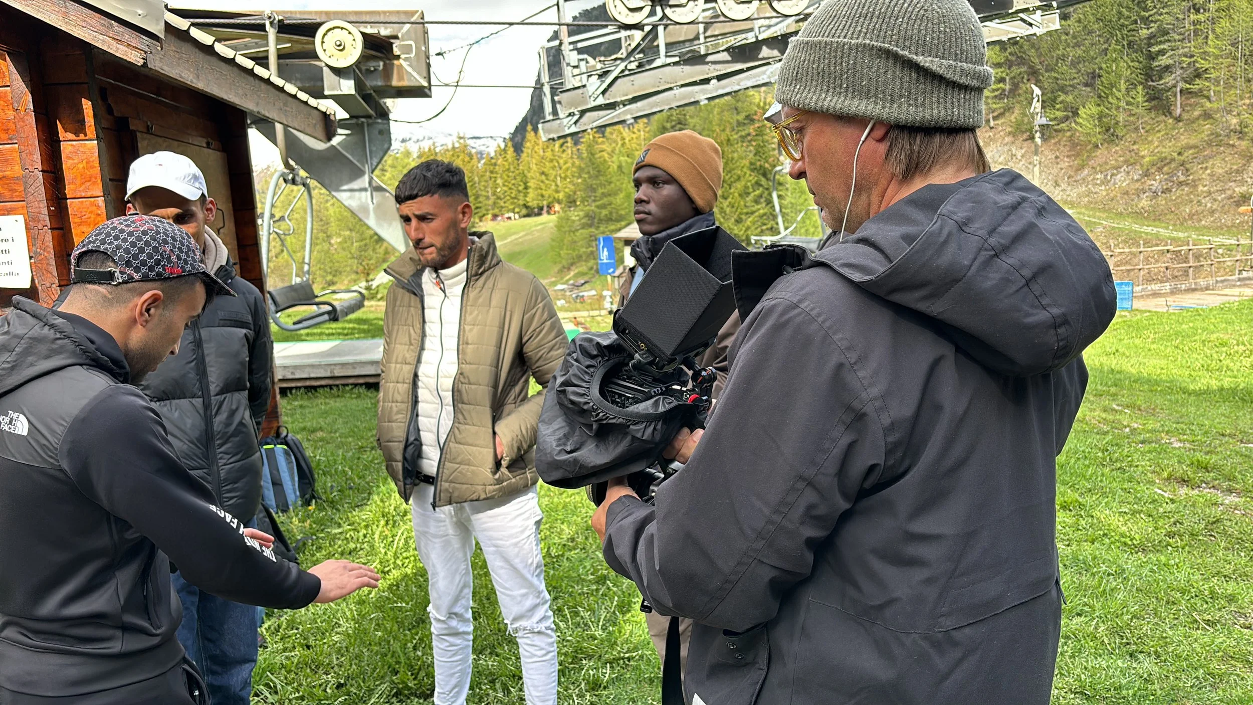 A group of five men outdoors in front of a ski lift, with three of them looking at a smartphone or device, while one is filming with a professional camera. The men are dressed in casual, warm clothing, and there are trees and a grassy area in the bac