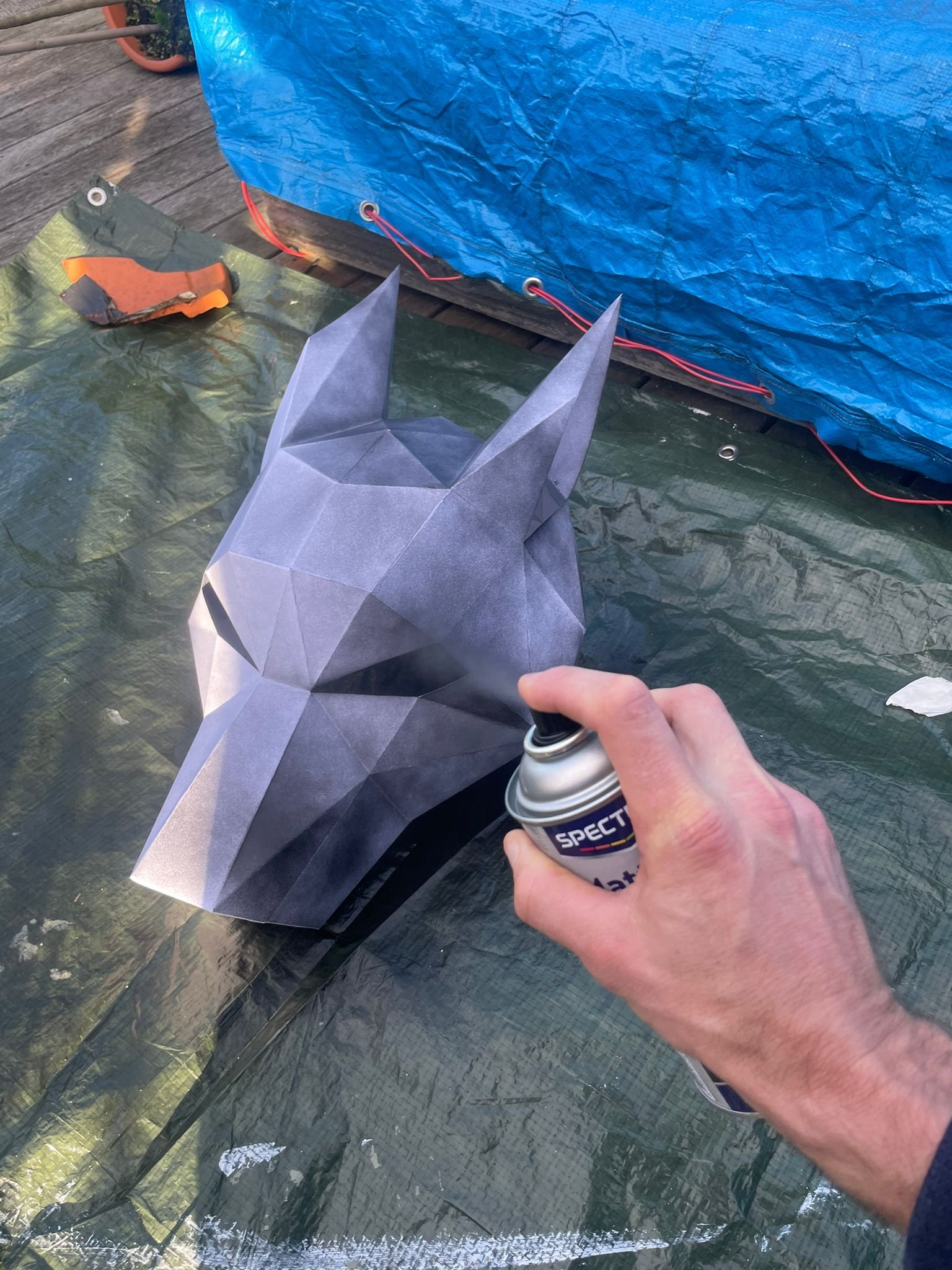 Person spraying a gray paper origami mask resembling a wolf or dog head with a can of spray paint on a green tarp outdoors.