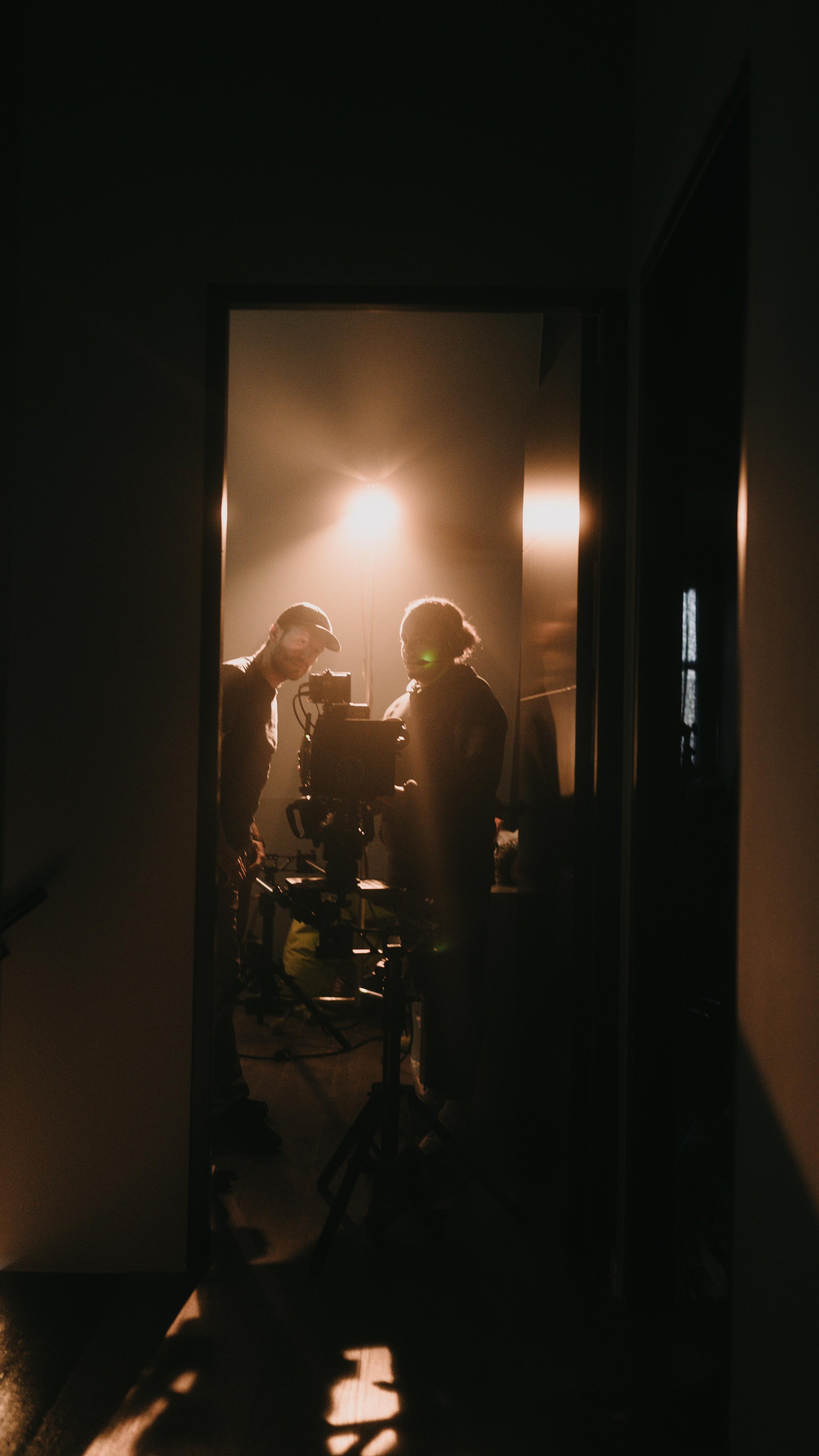 Silhouetted people working on a camera inside a dimly lit room, viewed through a doorway with backlighting.