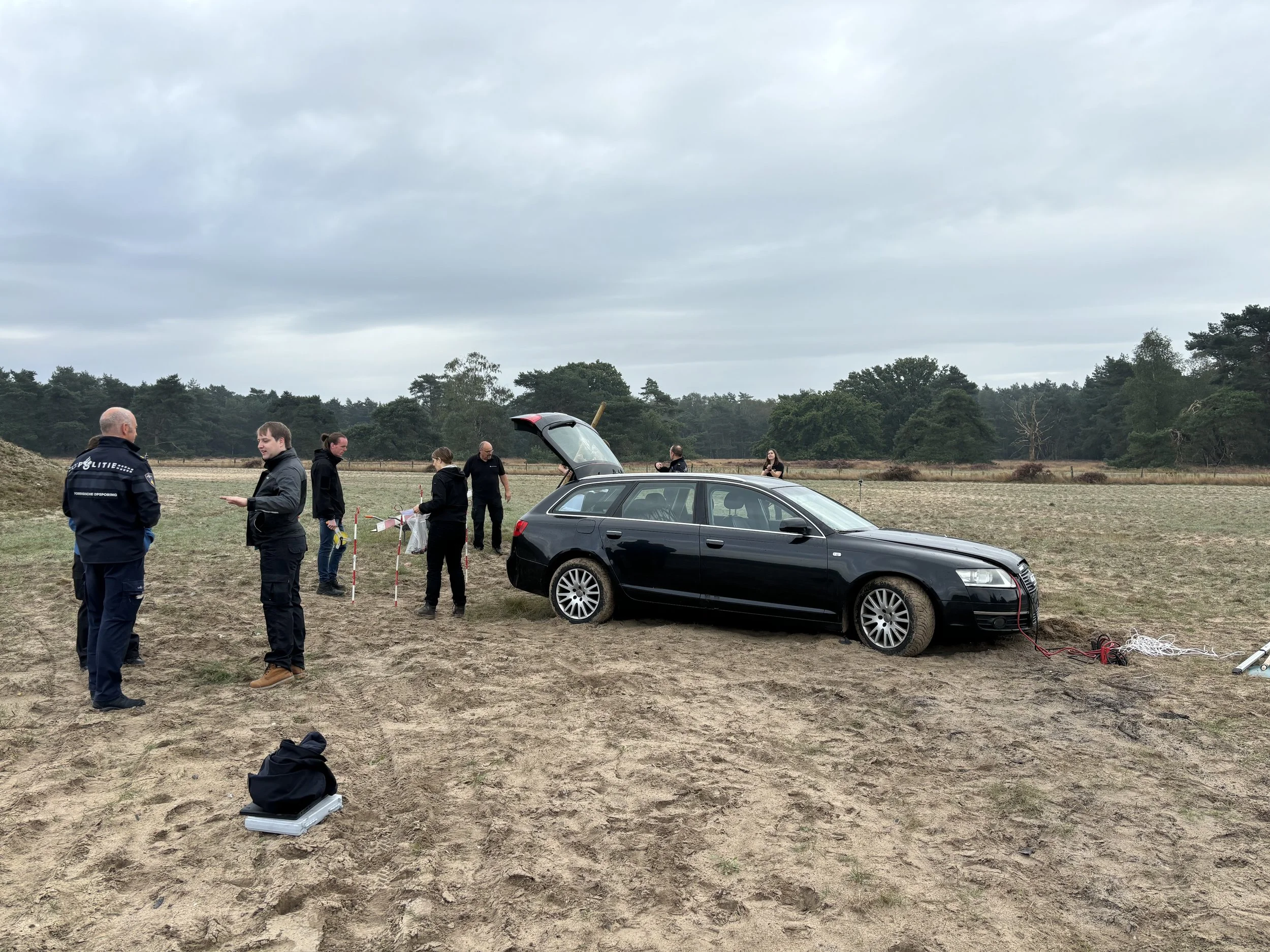 A black Audi car is off the road in a field with one wheel on the ground and the others possibly stuck, connected to a power source. Several people, including police officers, are gathered around the vehicle, some taking notes and others observing. T