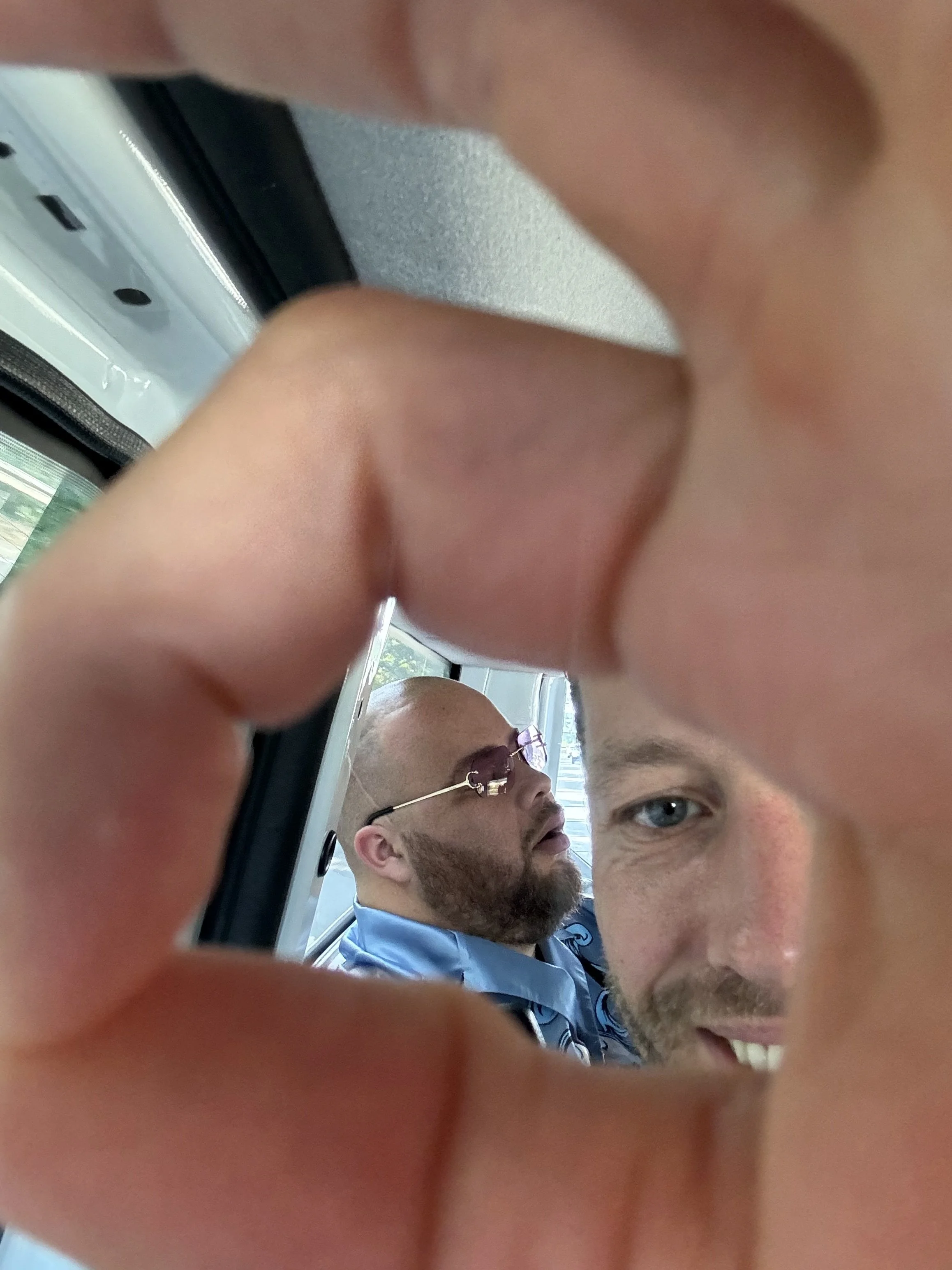 A person holds a device to take a photo through a small opening, capturing two men seated next to each other on a bus. One man has a beard, sunglasses, and is wearing a blue shirt; the other man is partially visible, smiling with a friendly expressio