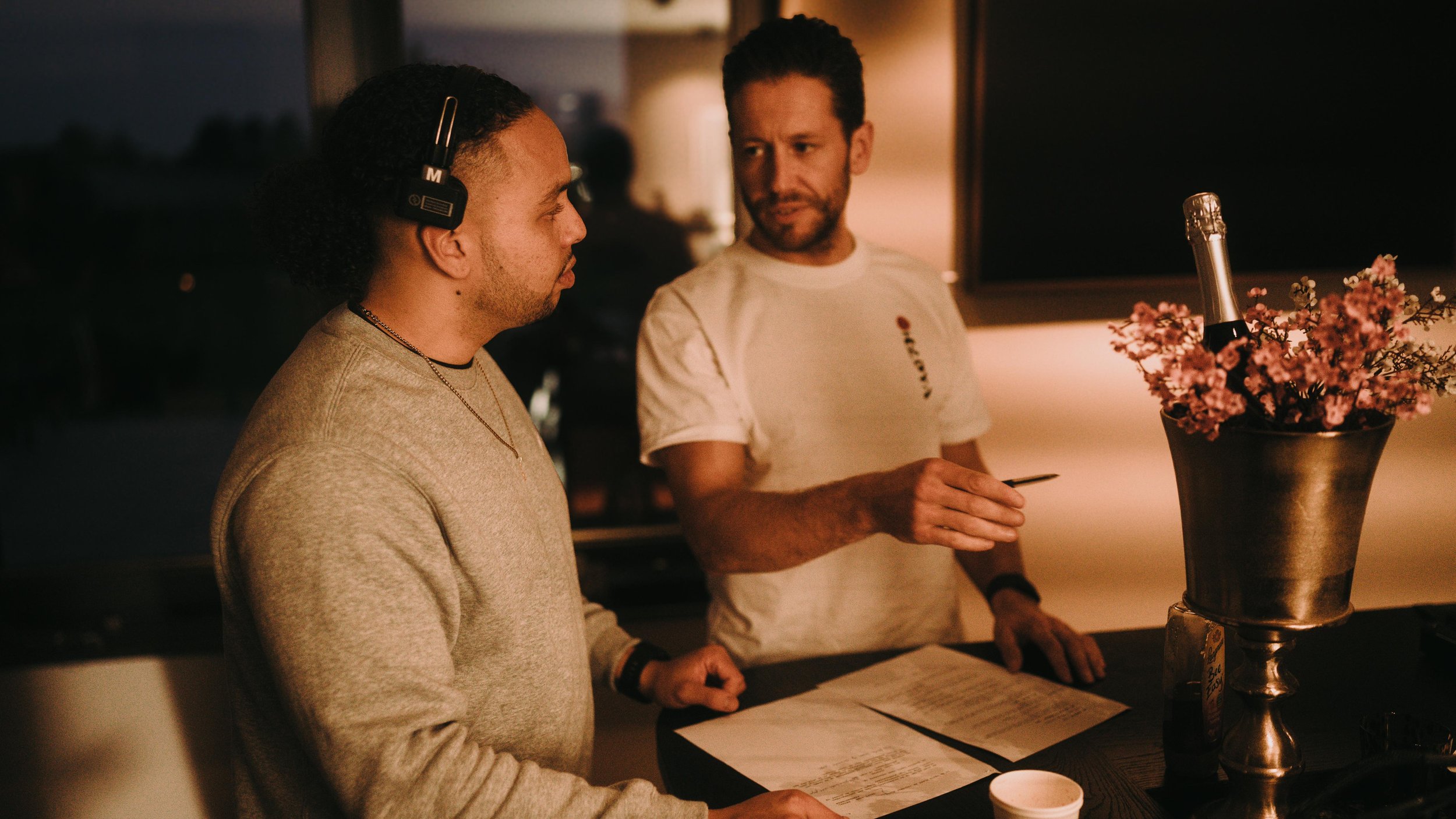 Two men having a conversation at a dimly lit bar or restaurant. One man is wearing a light-colored sweater and headphones, while the other is in a white t-shirt, holding a pen. There are papers and a flower arrangement with a champagne bottle on the 