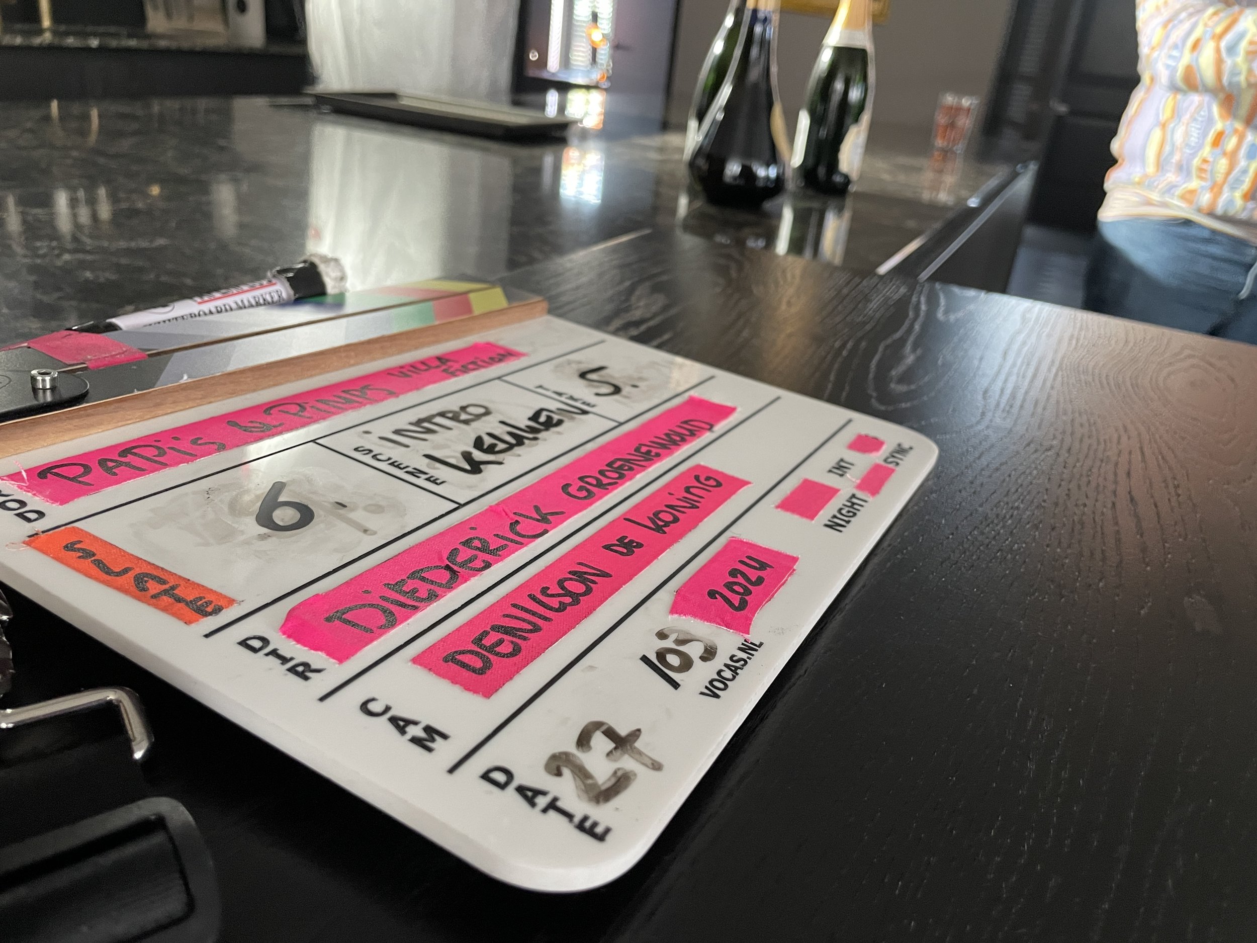 A clapperboard used for filming, placed on a dark wooden table, with bottles and part of a person's body in the background.