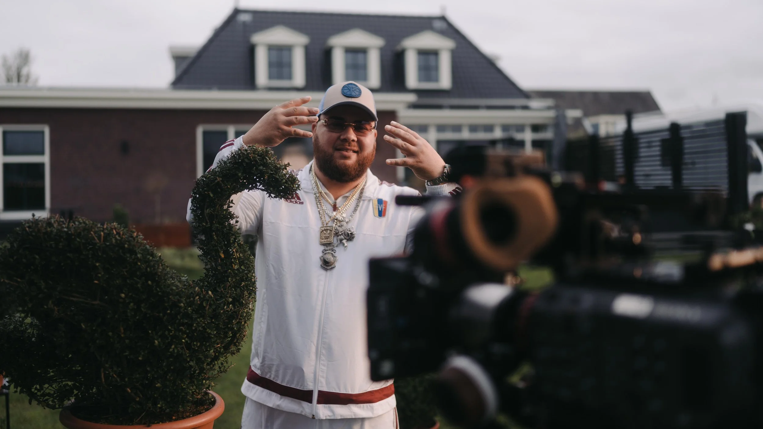 A man with a beard and sunglasses poses outdoors in front of a house, wearing a white jacket with patches, gold chains, and a cap, while facing a camera.