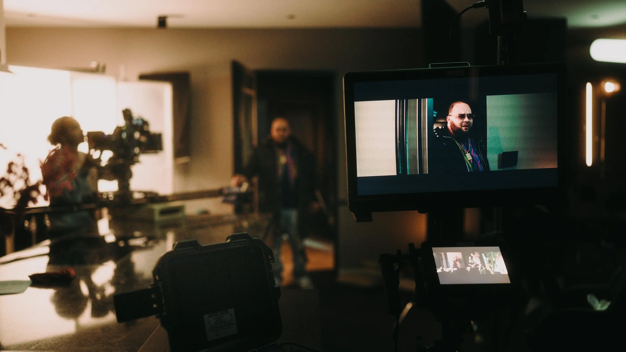 A filming scene showing a man with sunglasses on a monitor, a woman with a camera, and a man walking in the background inside a dimly lit room.