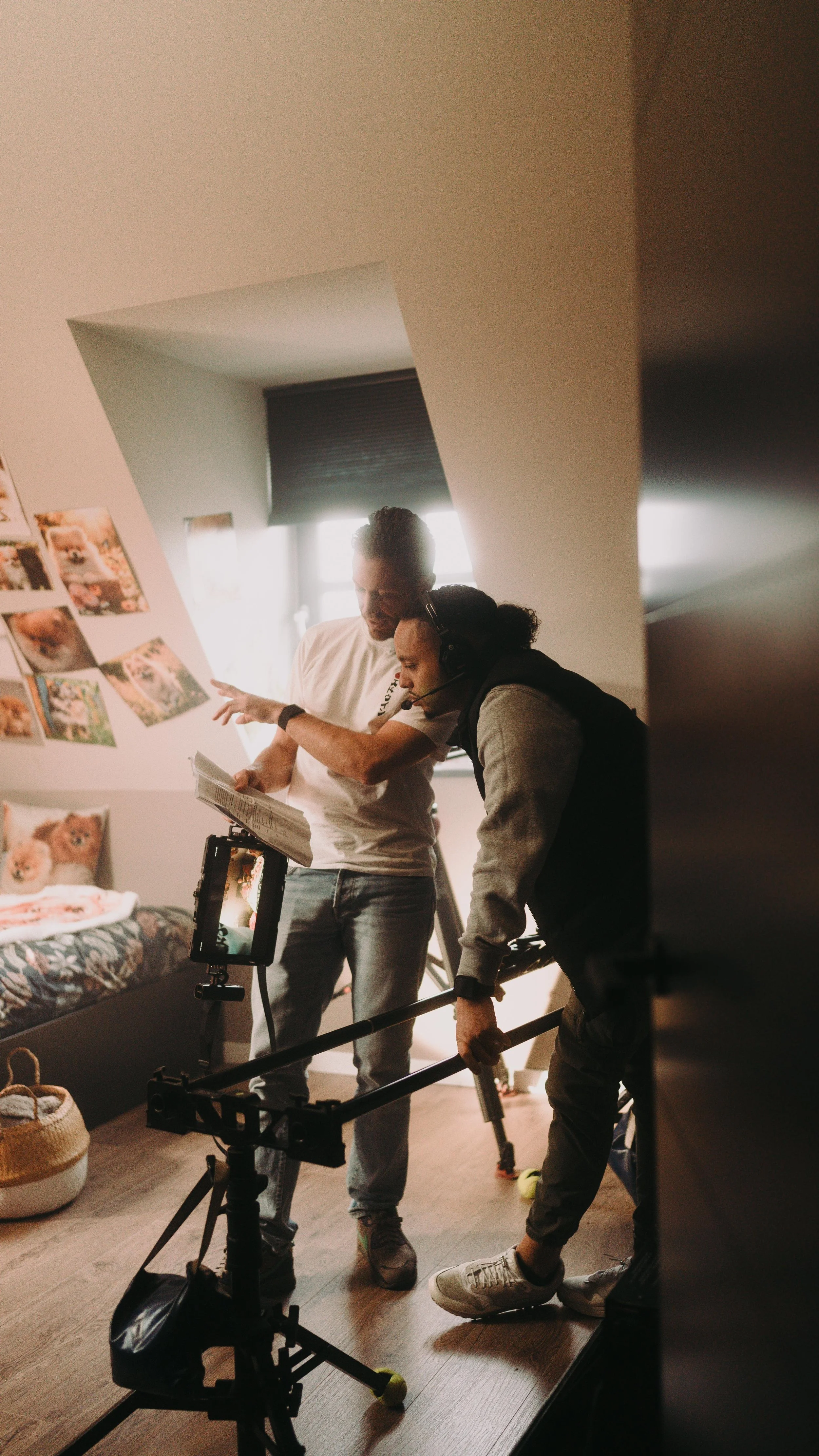 Two men working on a film set inside a bedroom, with photos of dogs on the wall and a bed with dog-themed pillows.
