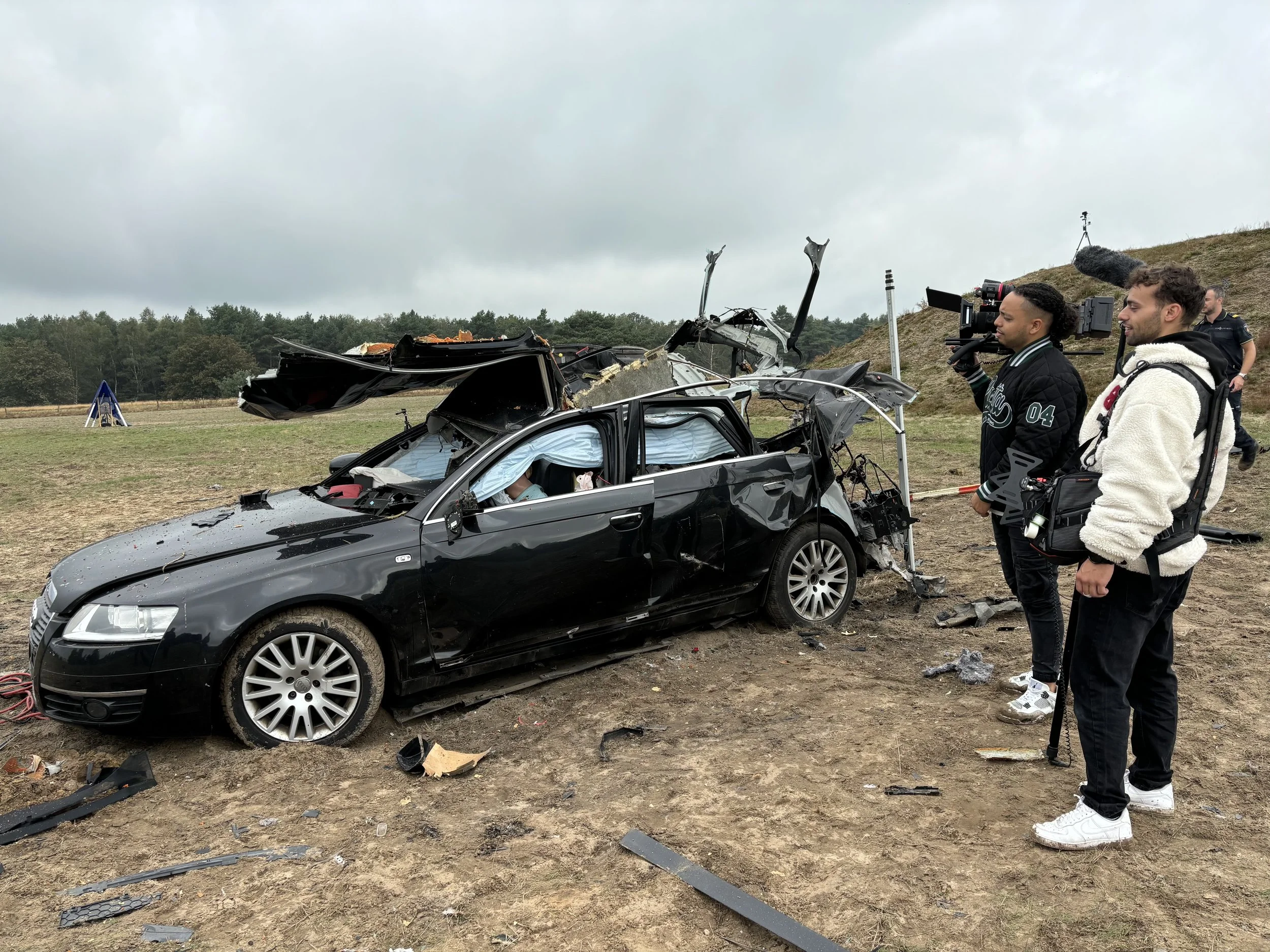 Camera crew filming a severely damaged black car after a crash on a field with overcast sky.