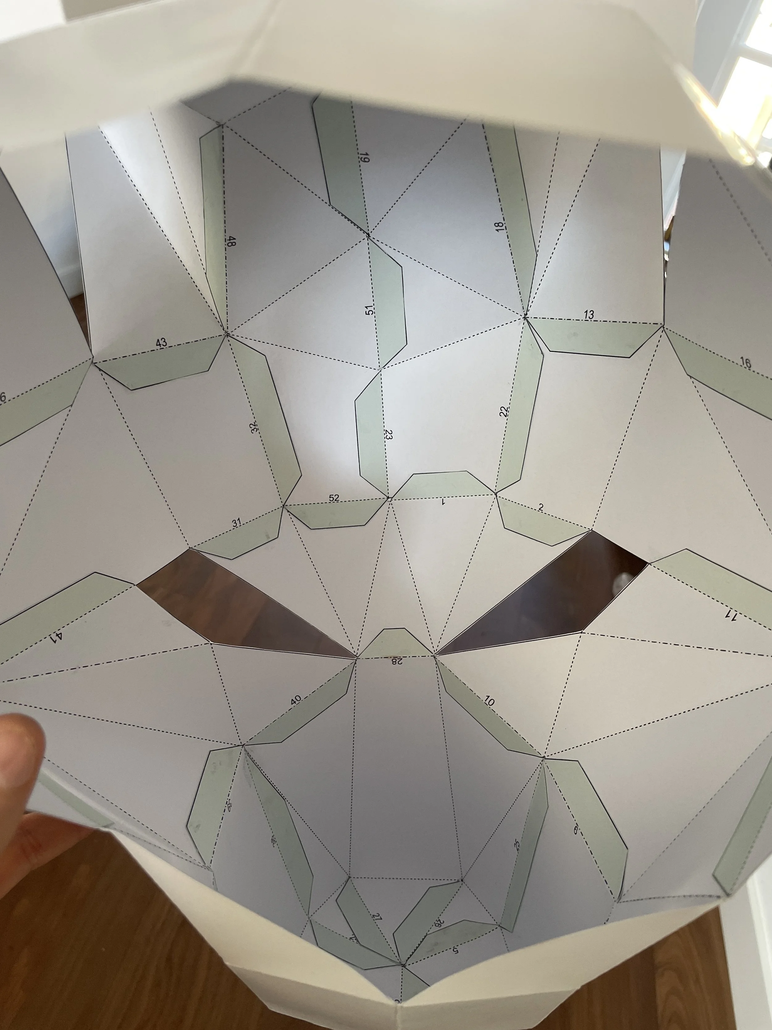 A person holding a paper mask template with cutouts for eyes and mouth, featuring numbered sections for assembly.