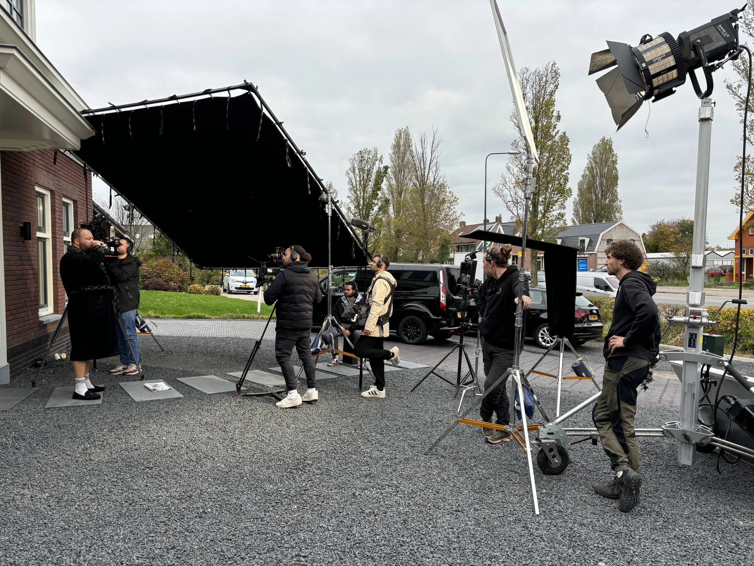 Outdoor film shoot with crew filming actors on a cobblestone driveway, large lighting equipment, and crew members wearing headphones and jackets, overcast sky, residential neighborhood background.