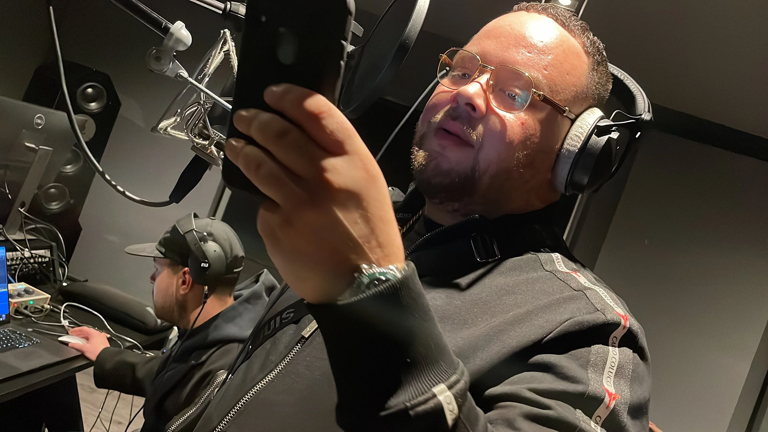 A man wearing headphones, glasses, and a dark jacket is recording vocals in a music studio. In the background, another person is working on a laptop with audio equipment.