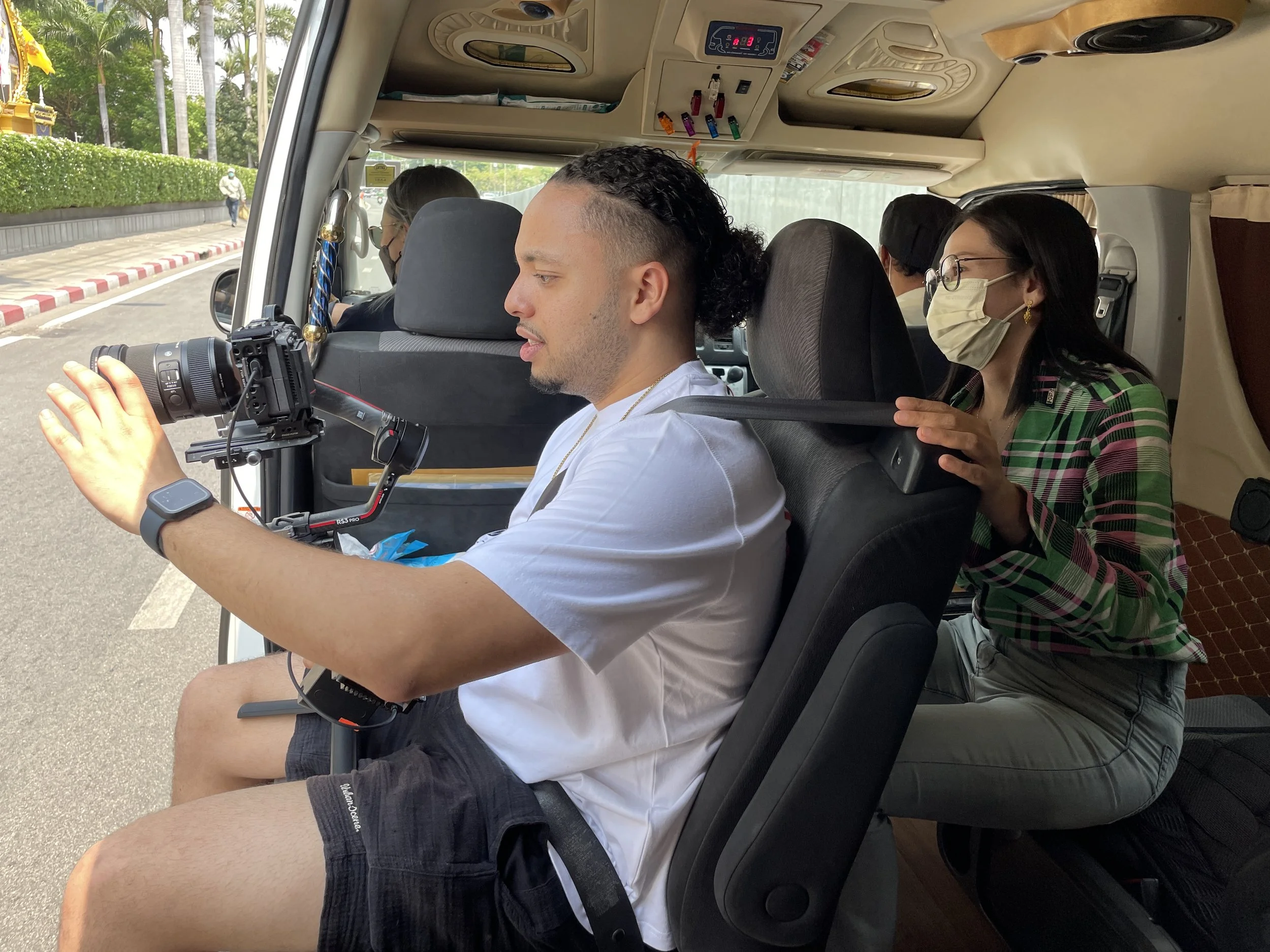 A man in a white t-shirt and shorts operating a camera mounted on a stabilizer inside a vehicle, with a woman behind him adjusting the seatbelt, wearing glasses and a face mask, and other passengers visible in the background.