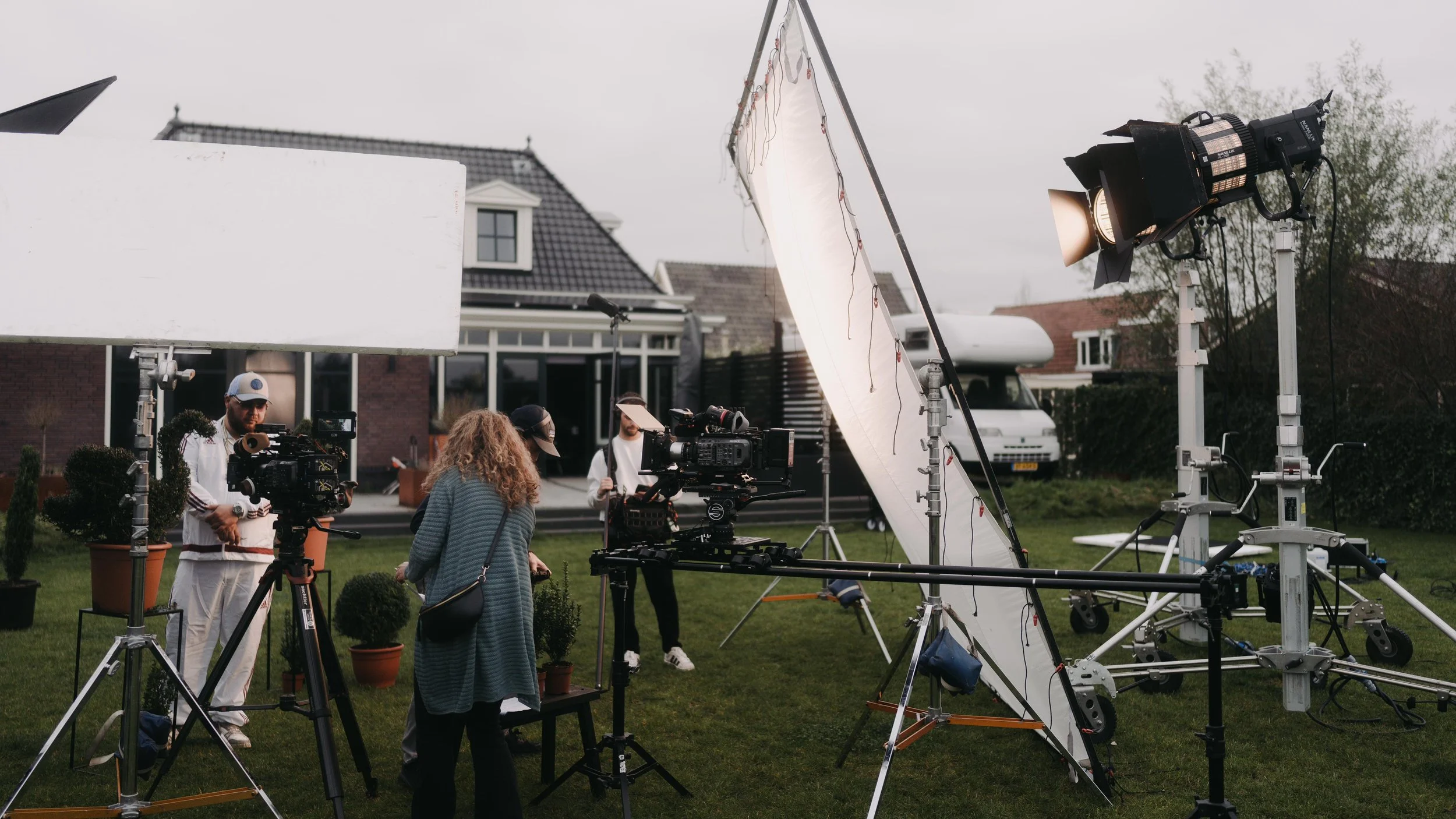 Film crew setting up outdoor shooting scene with professional cameras, large lighting, and reflectors in a garden outside a house.