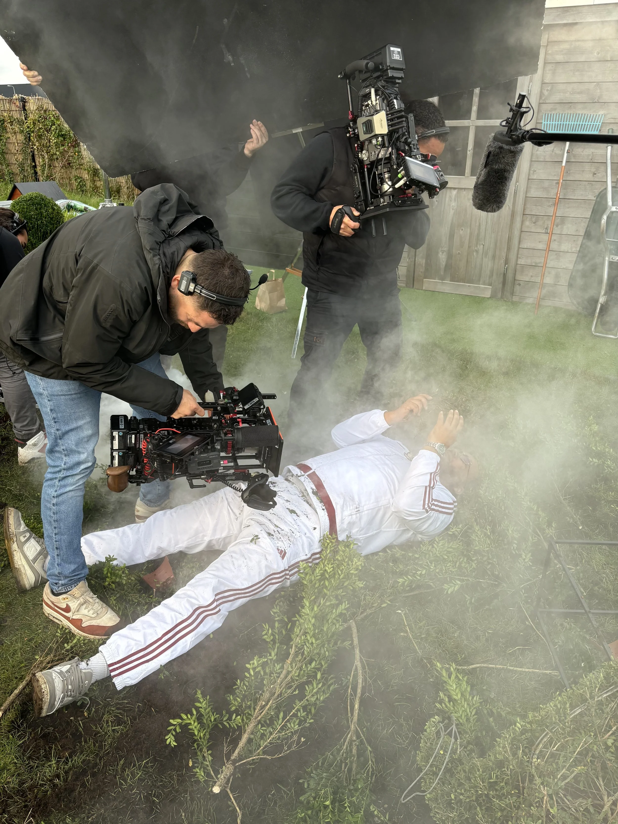Behind the scenes of a film production showing a person lying on the ground in white clothing, surrounded by filming crew and camera equipment, with smoke and foliage nearby.