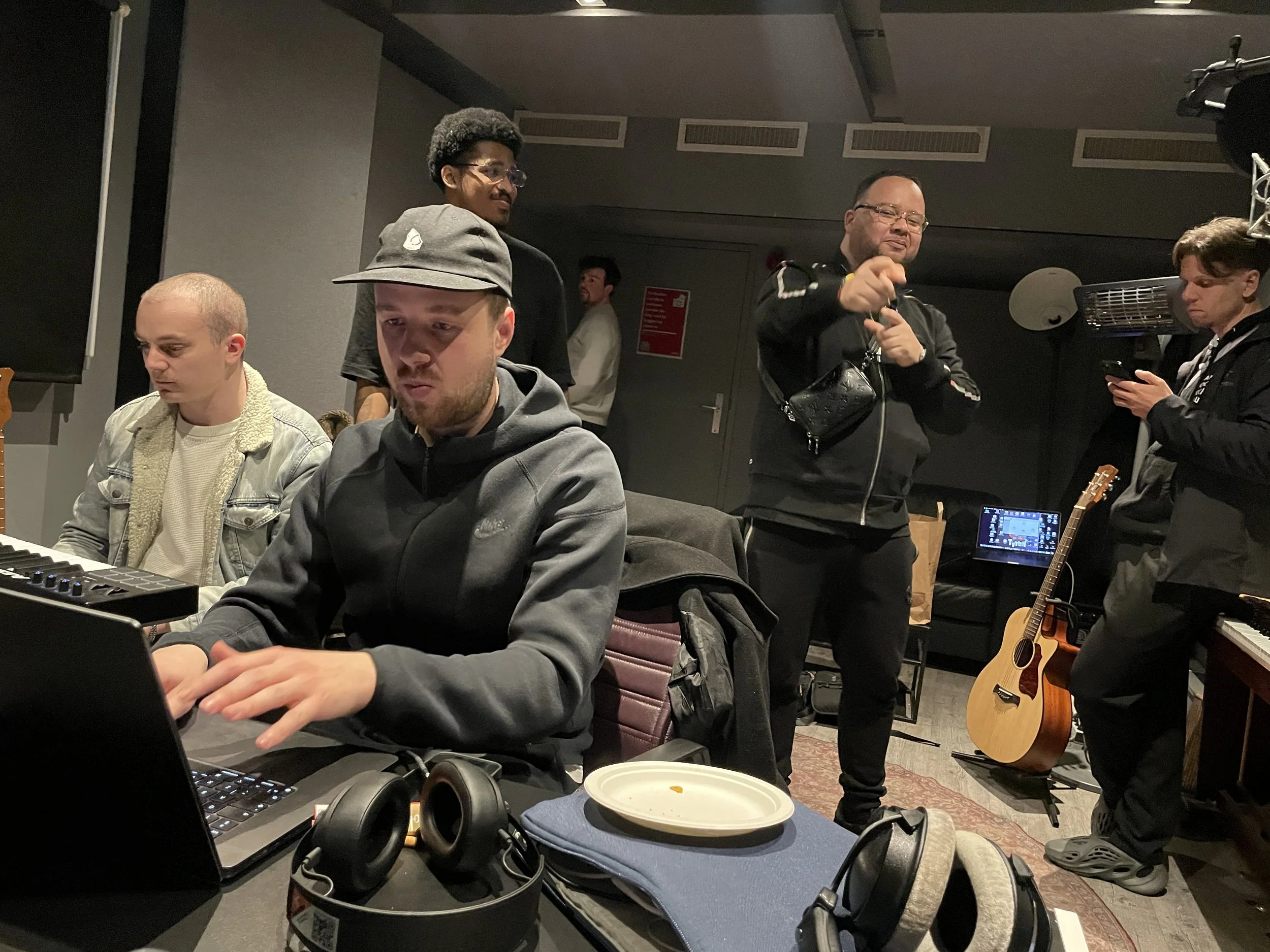 Group of people engaged in music production in a studio, with two individuals seated at a table with laptops and music equipment and three standing around, one with a guitar.