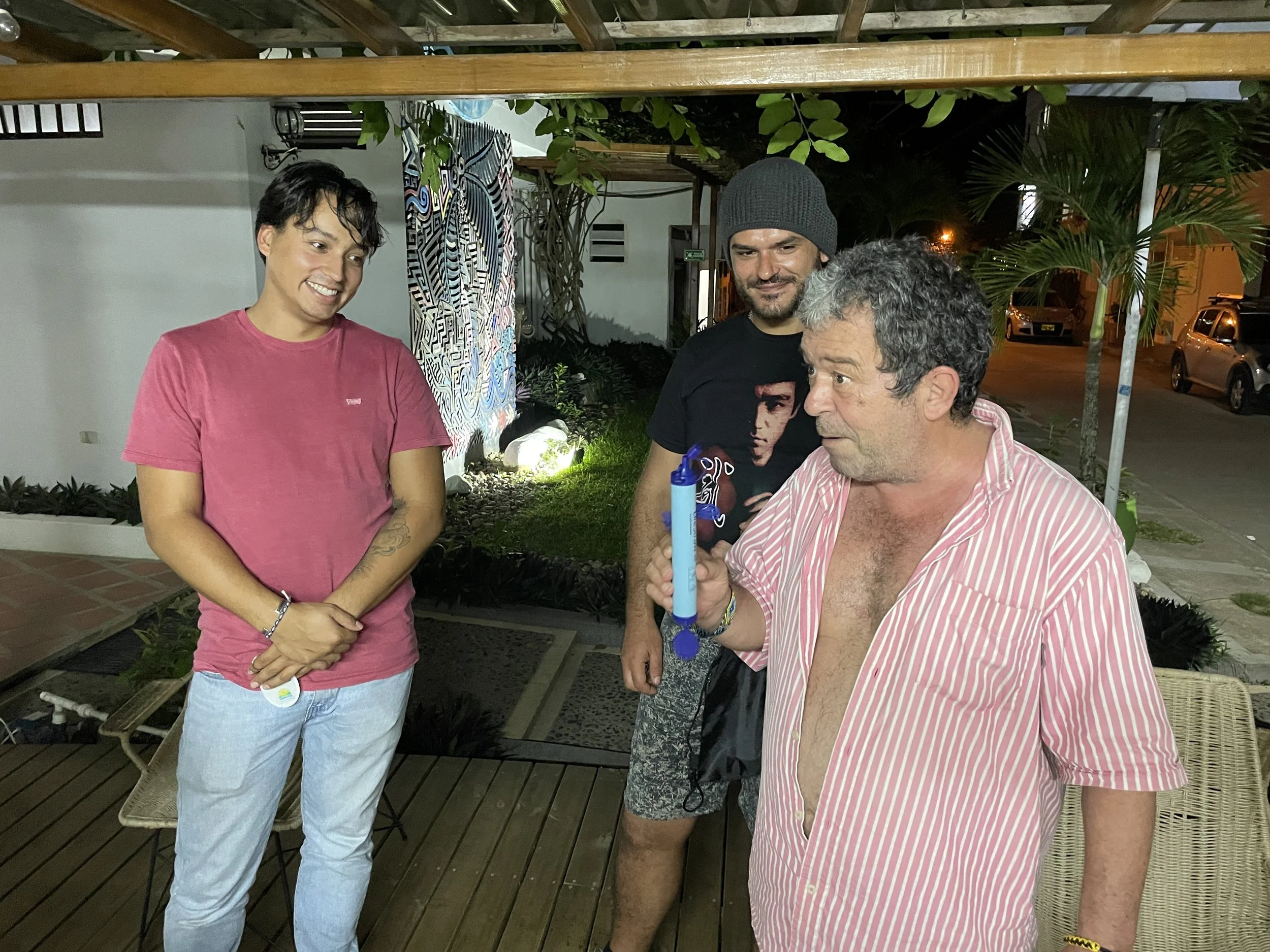 Three men are standing outside at night, one of them is holding a blue object and appears to be blowing into it, while the other two are watching and smiling.