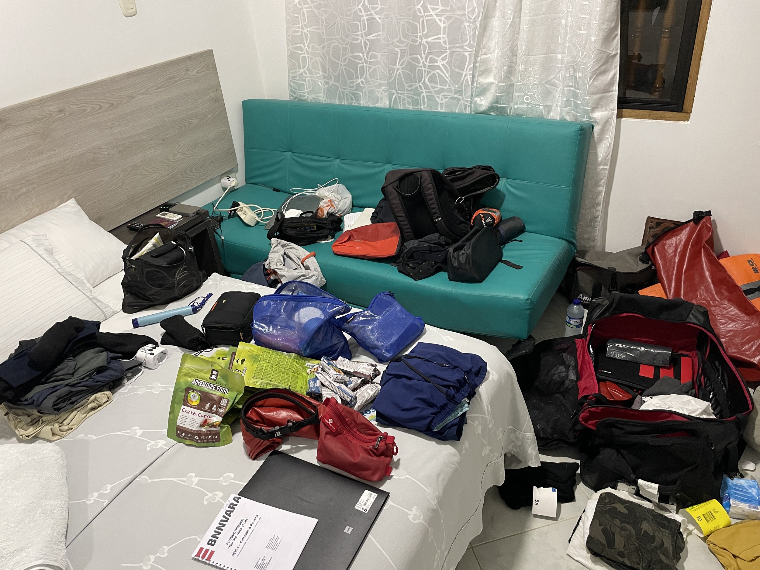 Hotel room with two beds and various packed bags, clothes, and supplies spread across the beds and floor, indicating someone is preparing for travel or a trip.