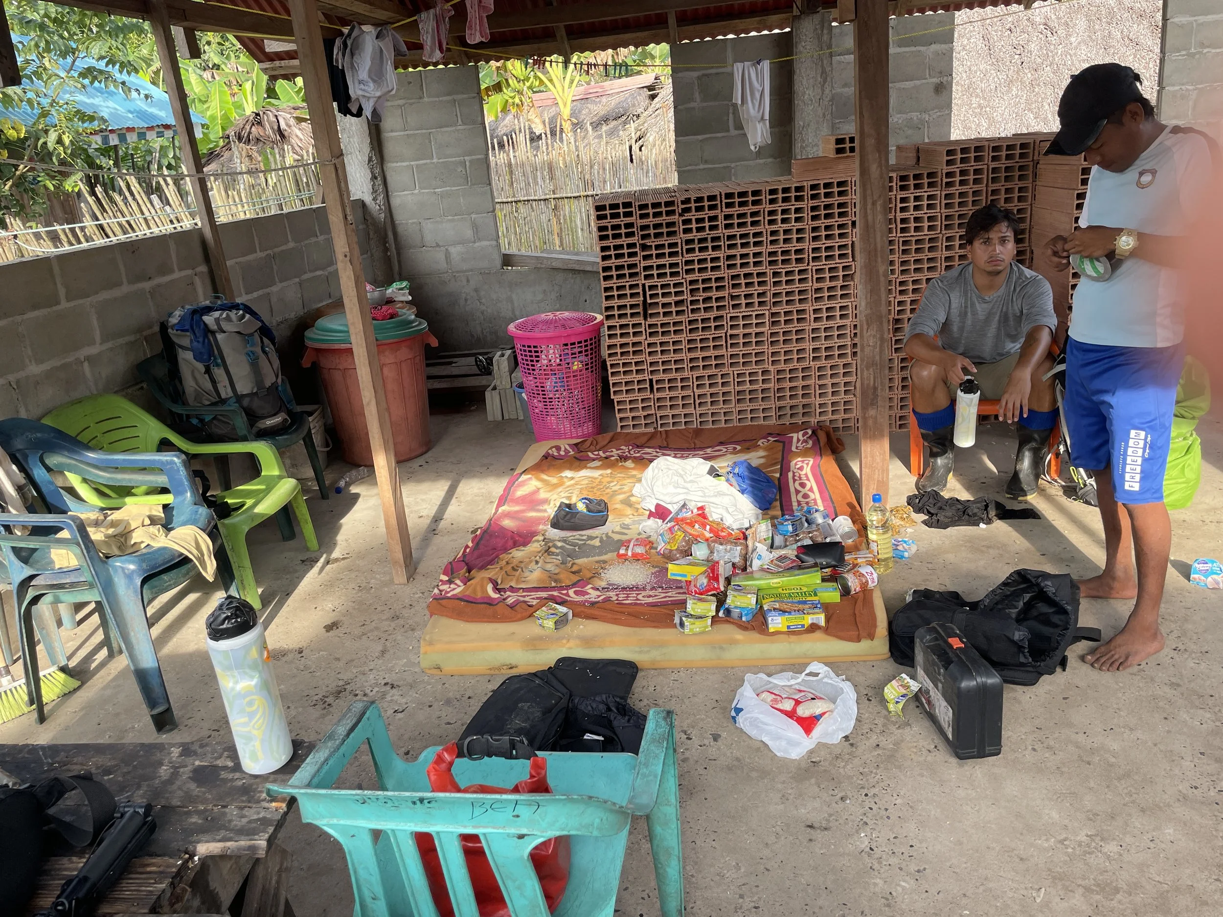 Two men are inside an unfinished shelter with a concrete floor and brick walls. One man is sitting on a small orange chair, holding a water bottle, while the other is standing, looking at some items in his hands. There are two piles of bricks behind 