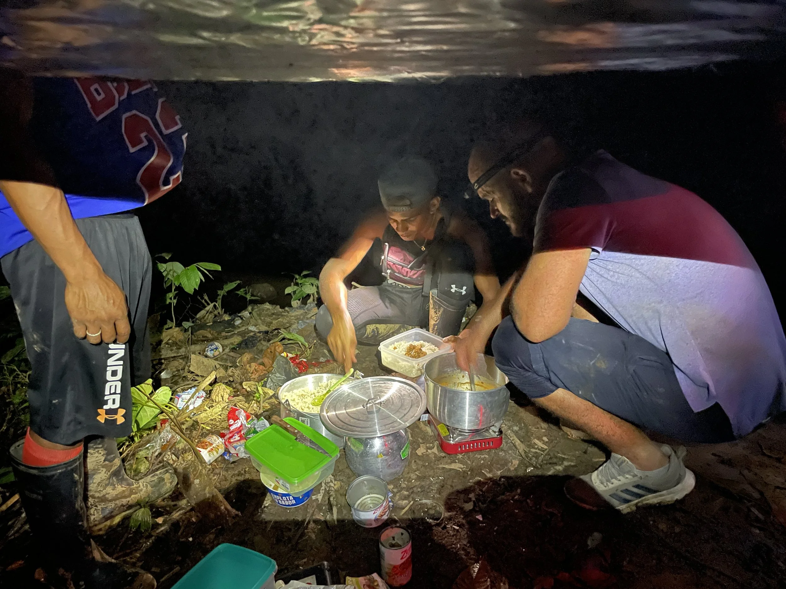 Three people preparing food under a rocky overhang at night with various cooking supplies and food containers on the ground.
