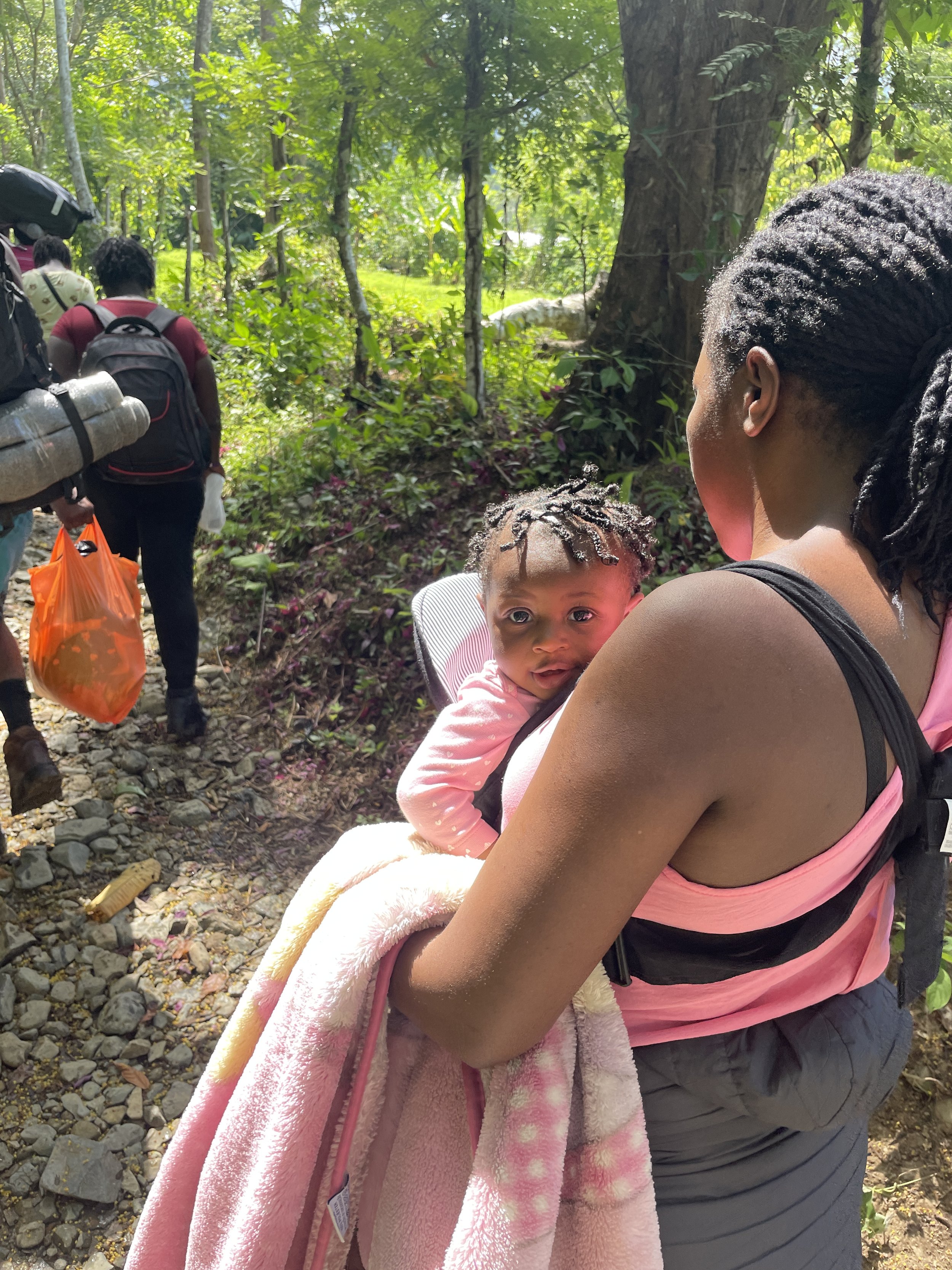 A woman holding a young child in a pink baby carrier on her back, walking on a forest trail with other hikers and lush green trees and plants in the background.